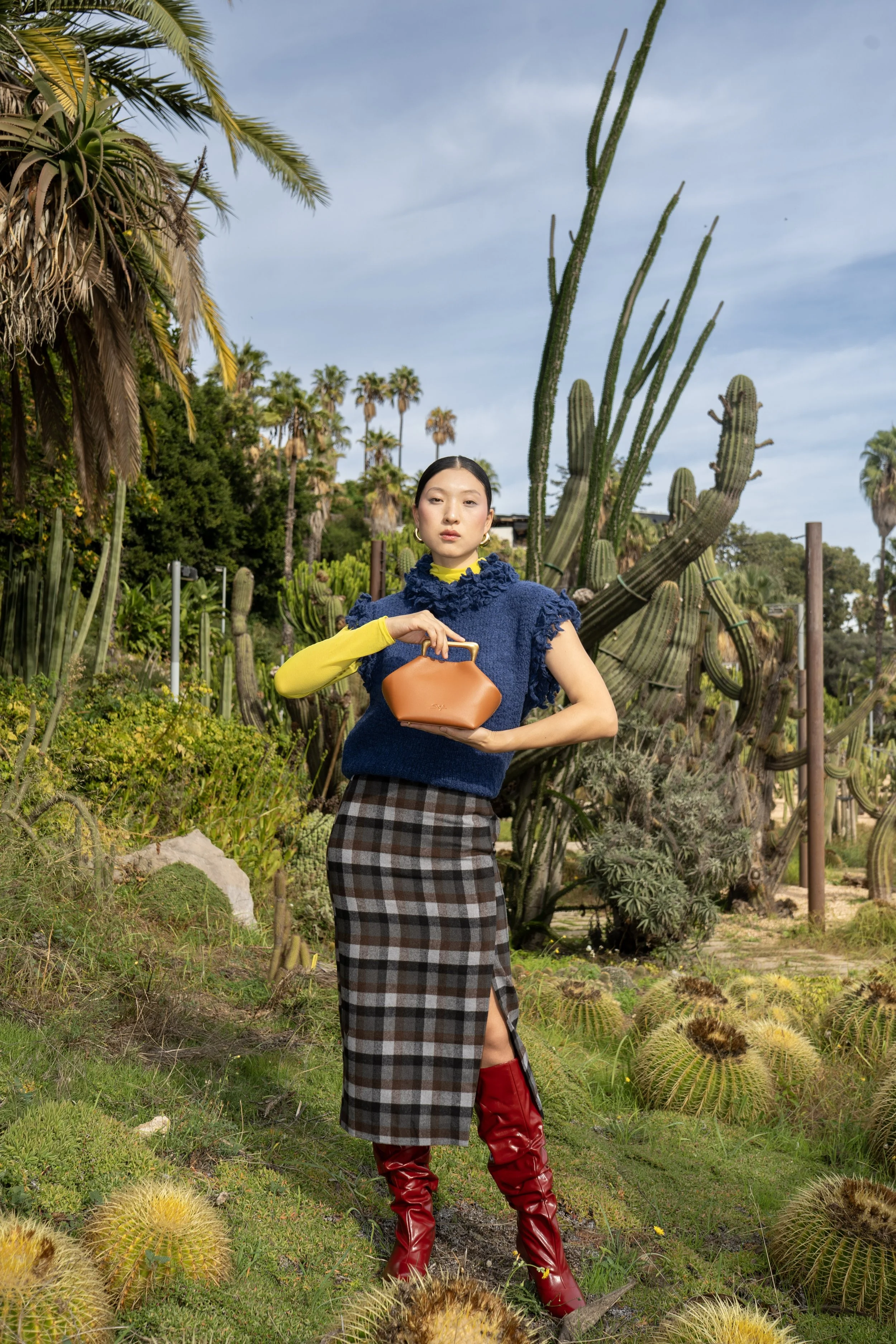 A woman standing in a desert garden with tall cacti and succulents, wearing a blue textured top, plaid skirt, yellow long-sleeve shirt, red boots, and holding a tan handbag.