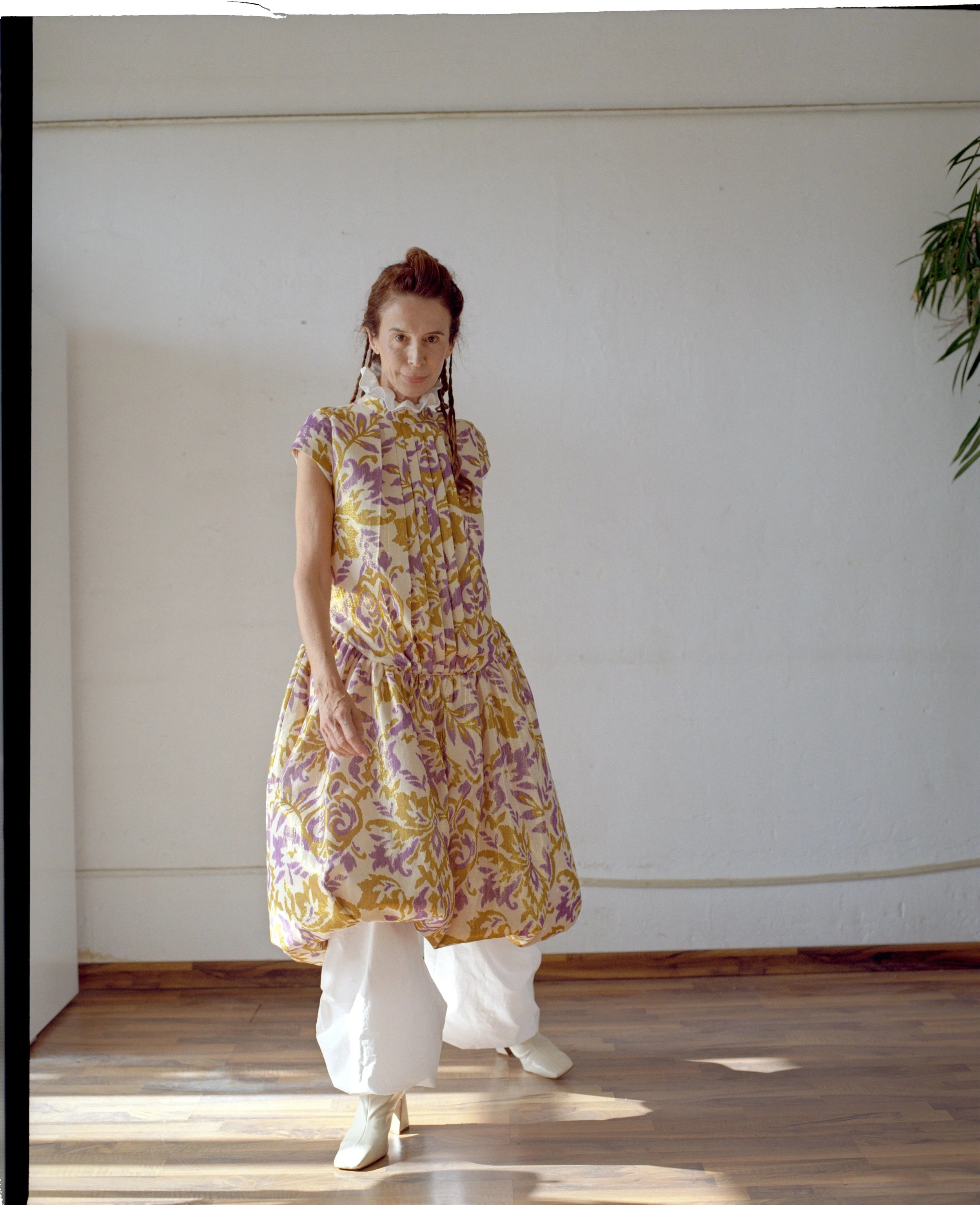 A woman standing indoors on a wooden floor, wearing a colorful, patterned dress with white, beige, and purple hues and white baggy pants with high-heeled white boots, with a white wall and a green plant partially visible on the side.