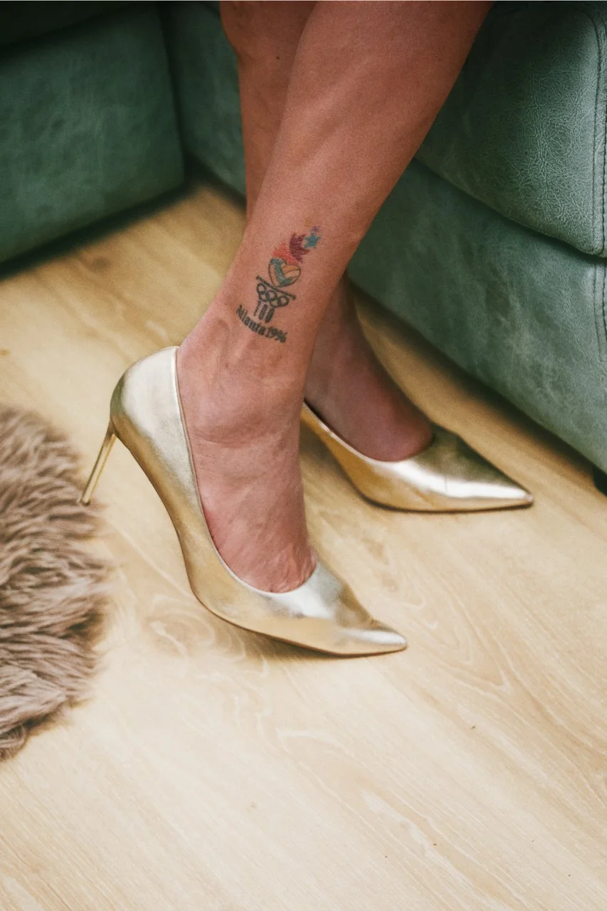 A woman's leg with a tattoo of a heart, cross, and angel wings on the ankle, wearing a gold high heel shoe.