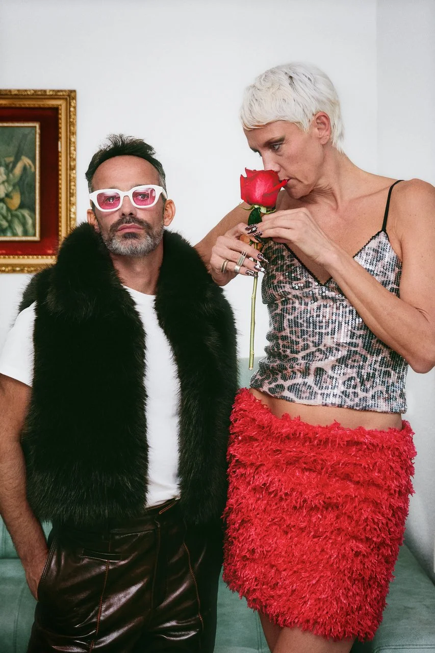 A man with dark hair, glasses, and a beard wearing a white shirt and black fur vest stands next to a woman with short platinum blonde hair, a sequined top, and a red feathered skirt. She is holding a red rose and appears to be smelling it.