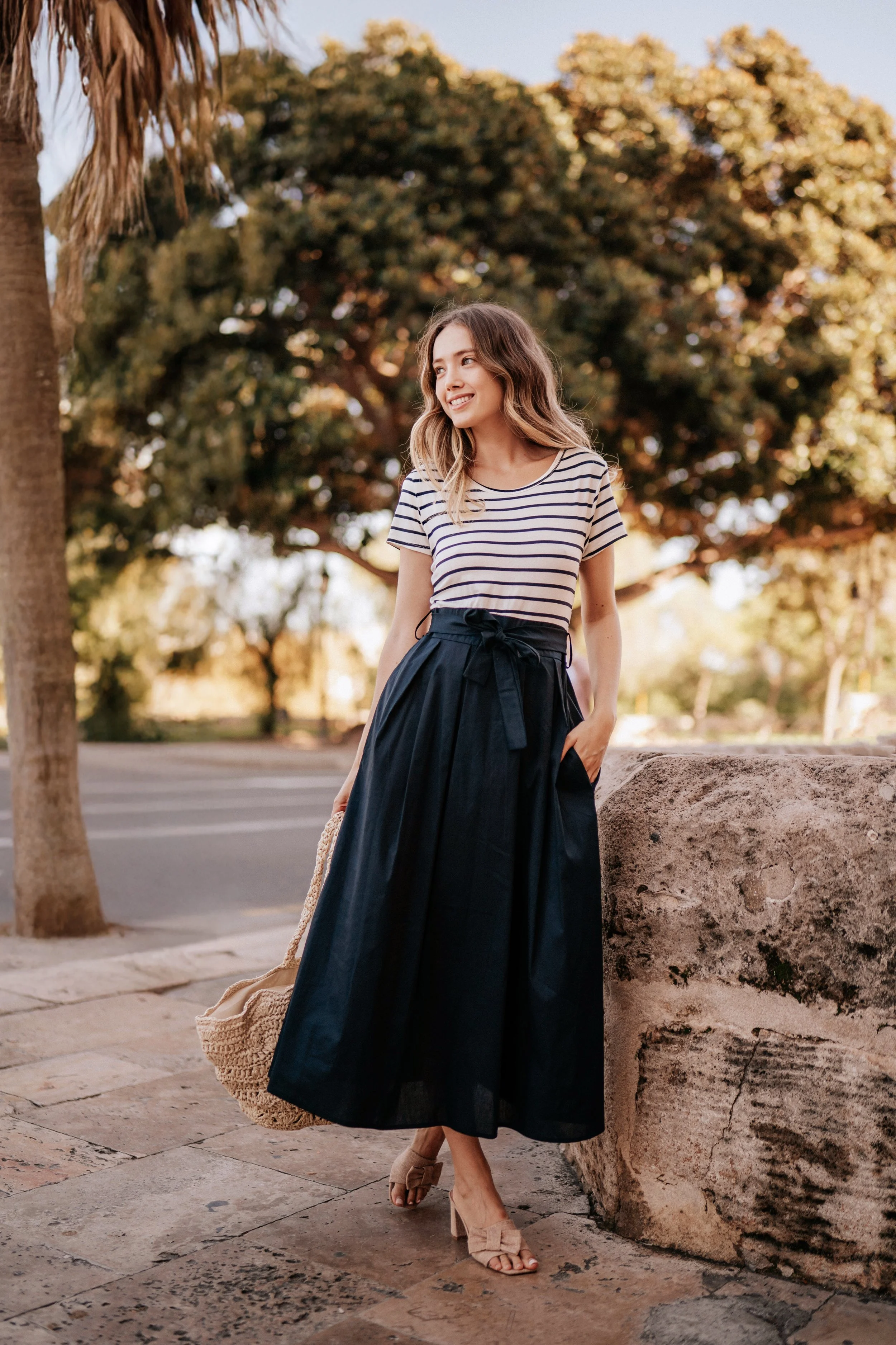 Young woman standing outdoors near a stone wall, wearing a striped t-shirt, navy skirt, and beige heels, holding a woven bag, smiling, with trees in the background.
