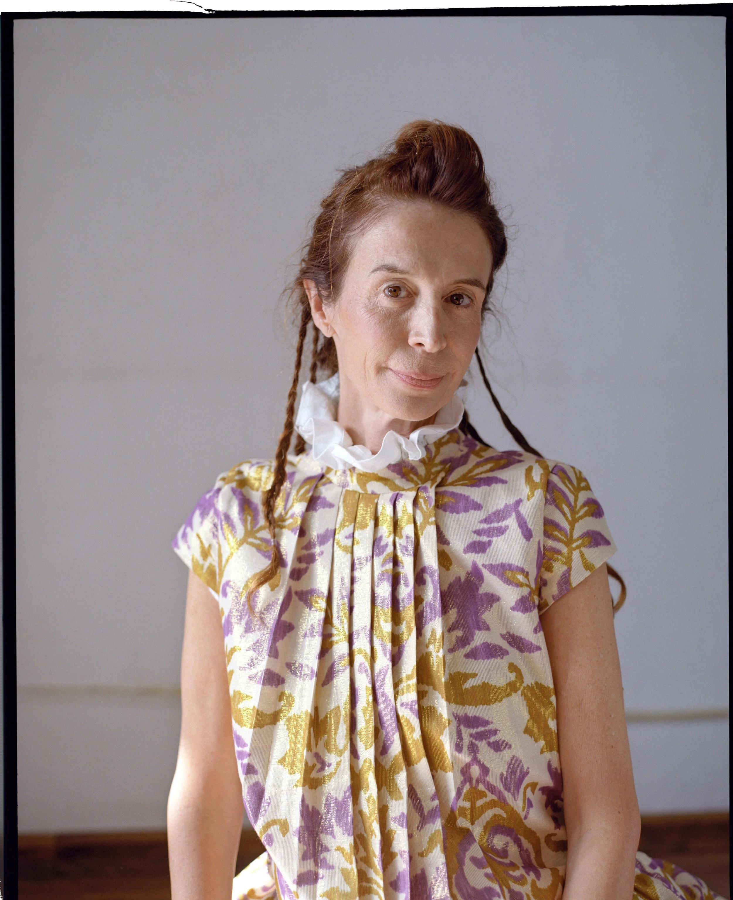 A woman with reddish-brown hair styled in a partial updo and braids, wearing a patterned dress with purple and gold leaves and a white ruffled collar, sitting against a neutral background.