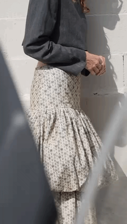 Person wearing a dark denim jacket and a beige floral ruffled skirt, standing against a white brick wall.