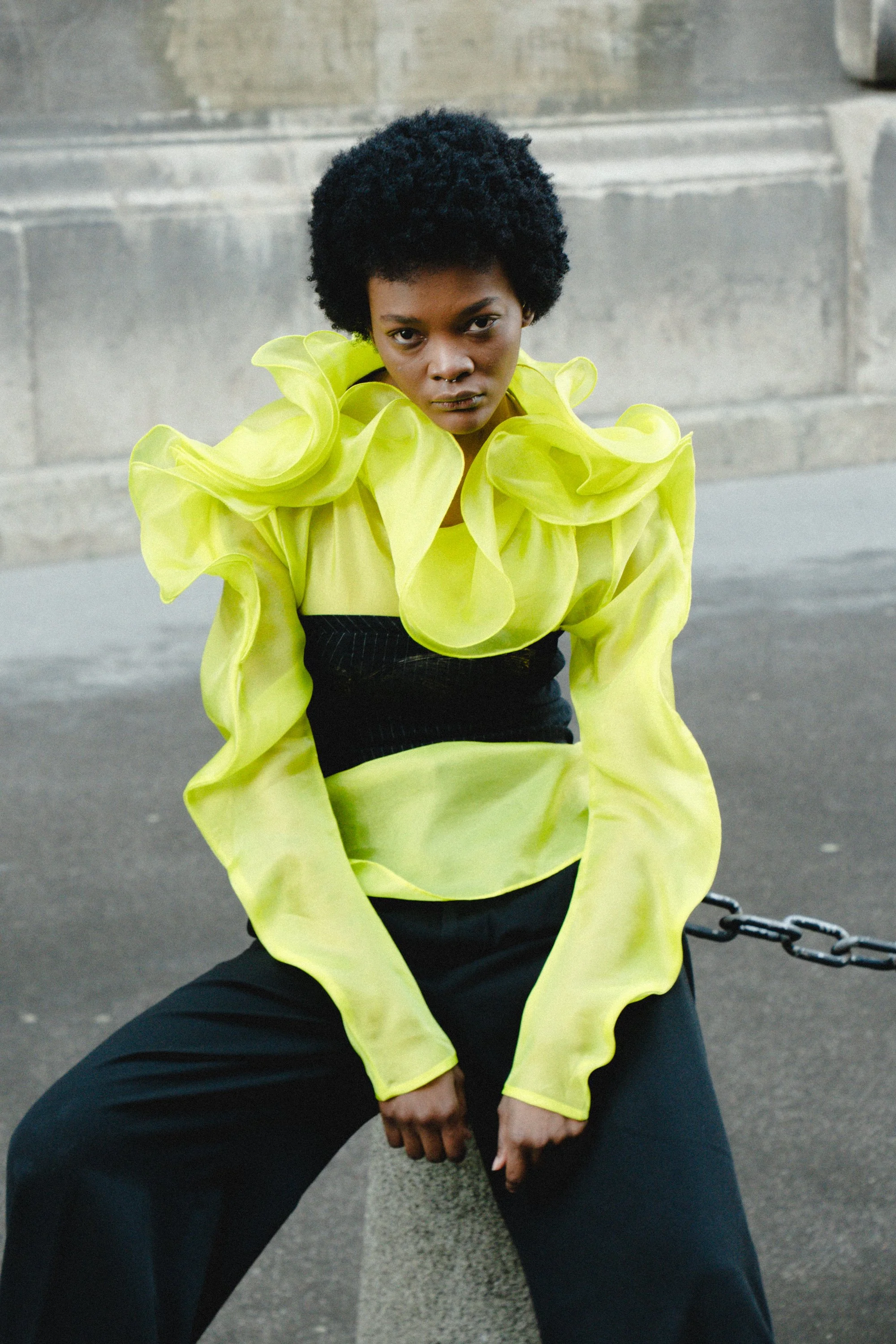 A woman with dark skin and styled curly hair poses outdoors wearing a bright yellow, ruffled top with long, sheer sleeves and black pants.