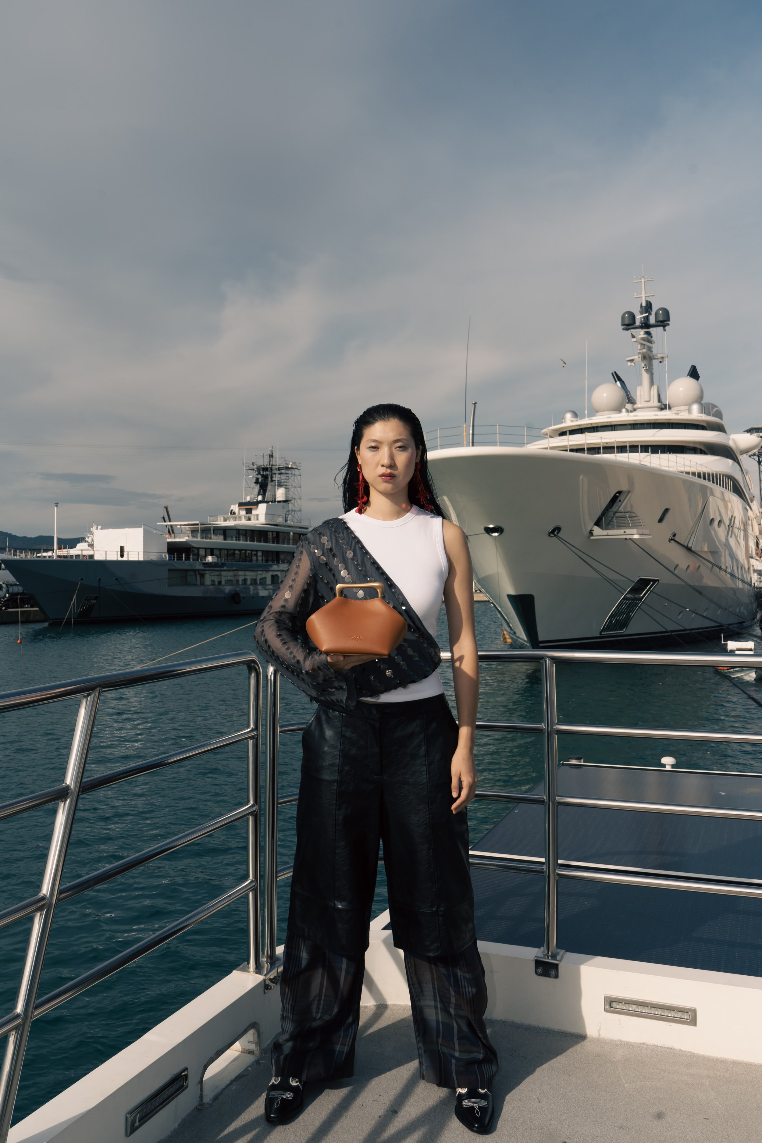A woman standing on a yacht dock with large yachts in the background, wearing a sleeveless white top, black leather pants, and holding a brown handbag.