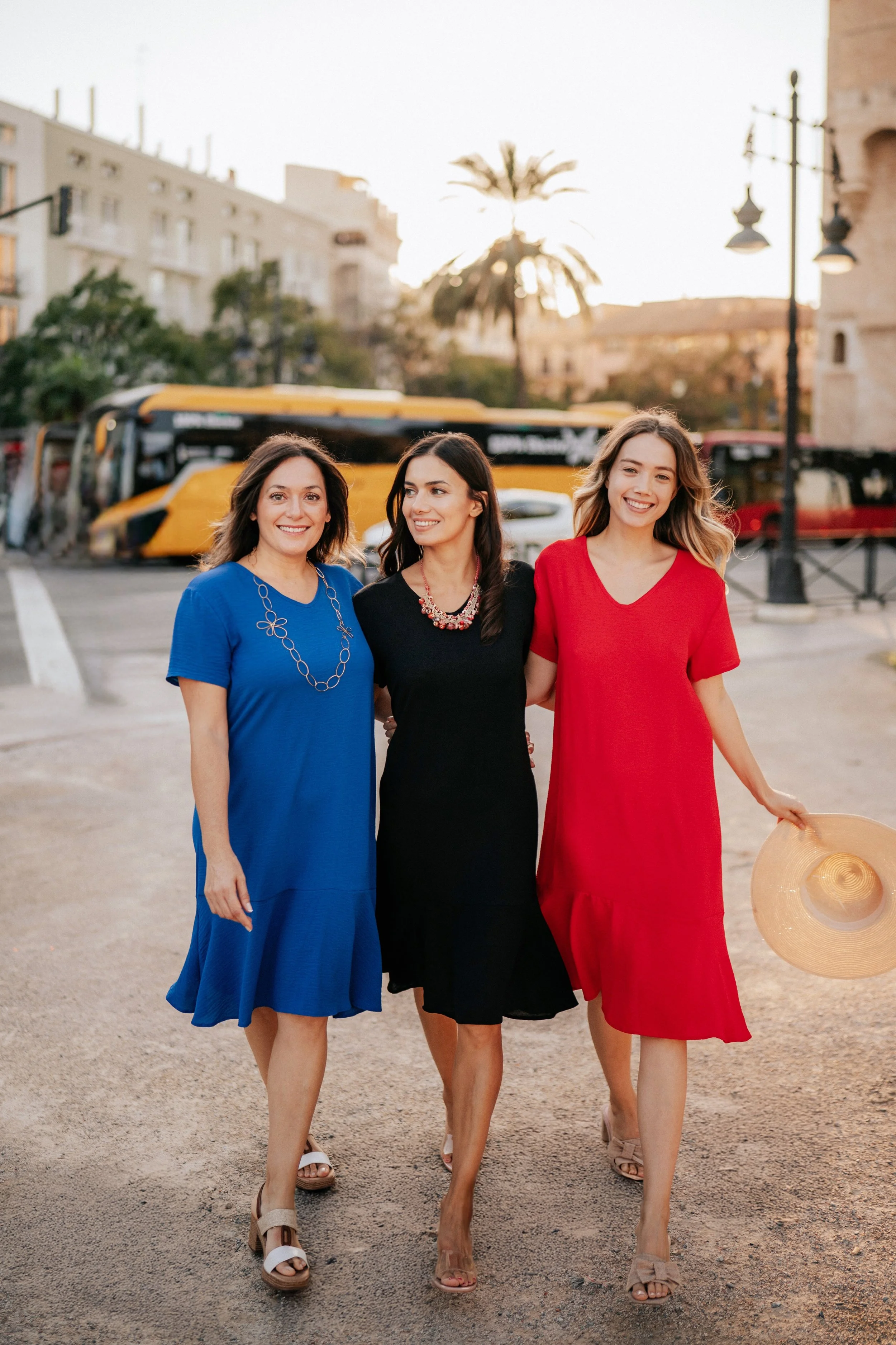 Three women walking arm-in-arm in a city street during sunset, smiling, with a yellow bus and palm trees in background, one woman holding a large straw hat.