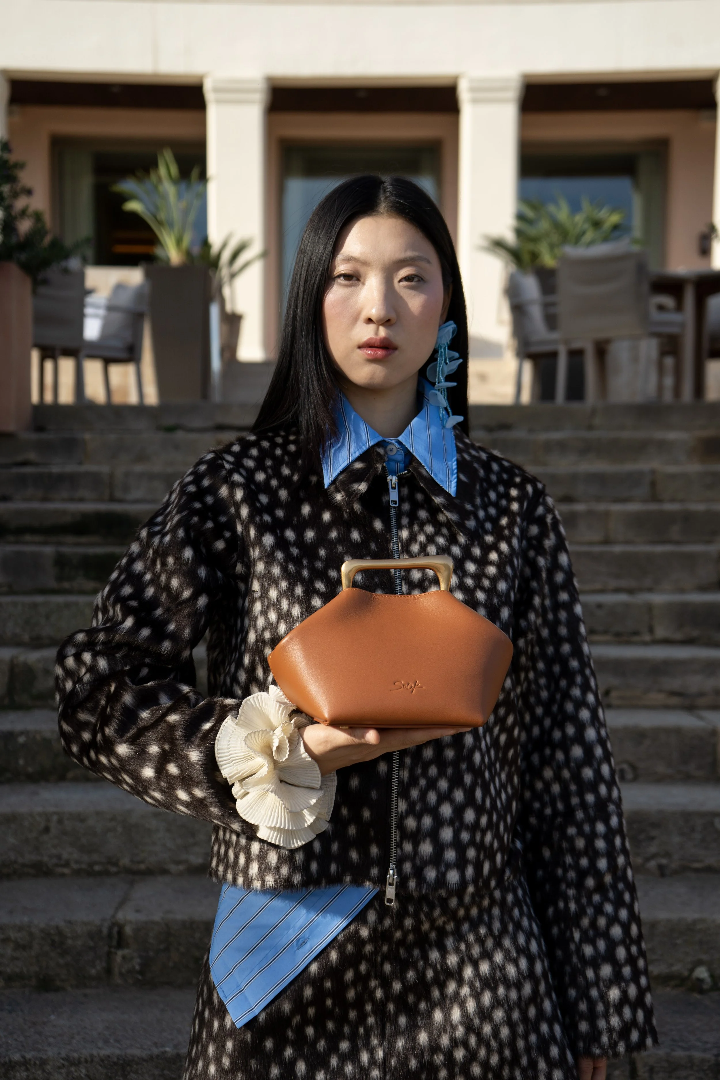 A woman with long black hair standing outdoors on stone steps, holding a small brown handbag, wearing a black and white patterned coat, blue striped shirt, and a beige scrunchie on her wrist.