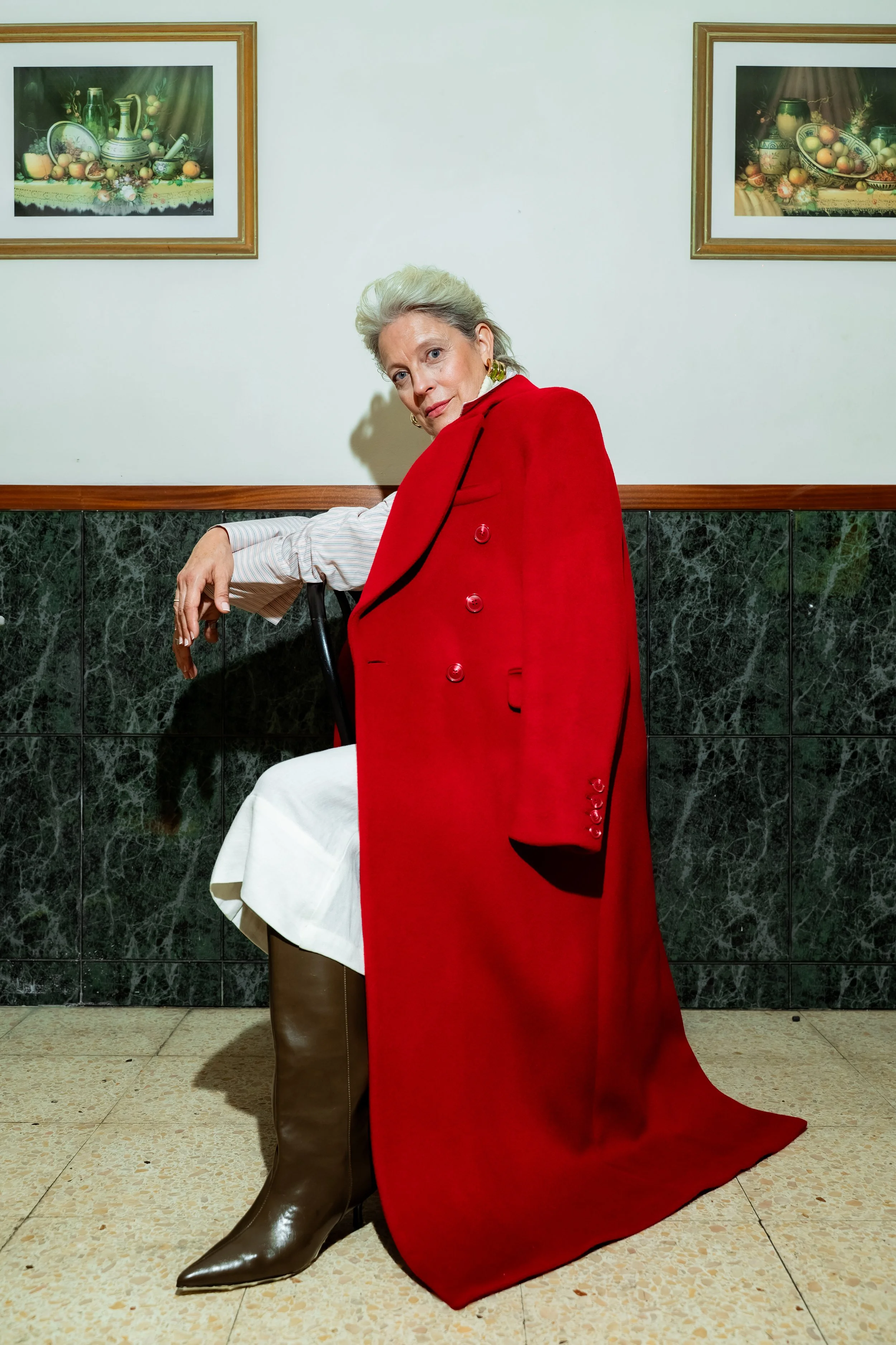 An older woman with gray hair and earrings sitting on a chair, dressed in a white skirt, brown knee-high boots, a white shirt, and a red cape, in a room with framed still-life paintings of fruit and kitchenware on the wall.