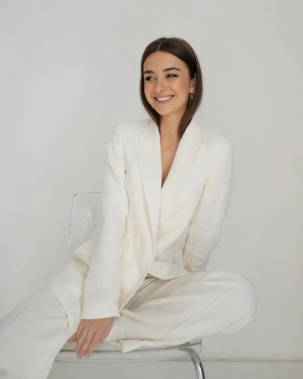 Cristina Marjinean - Founder of Verso Visual  - A young woman with dark brown hair and earrings, smiling and sitting cross-legged on a clear chair, wearing a white suit against a plain white background.