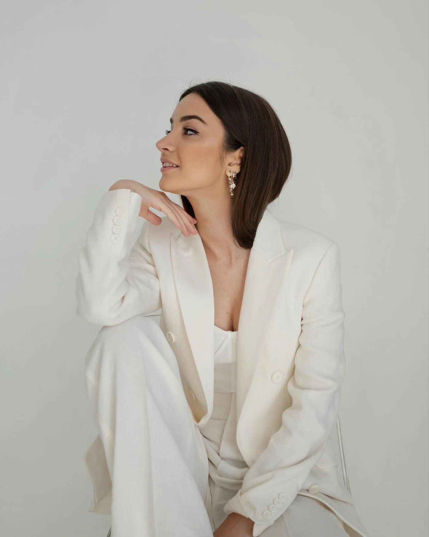 Cristina Marjinean - Founder of Verso Visual  - A woman with shoulder-length dark hair, wearing a white blazer and matching pants, is sitting against a plain light background, posing with her hand resting on her chin, looking to the side.