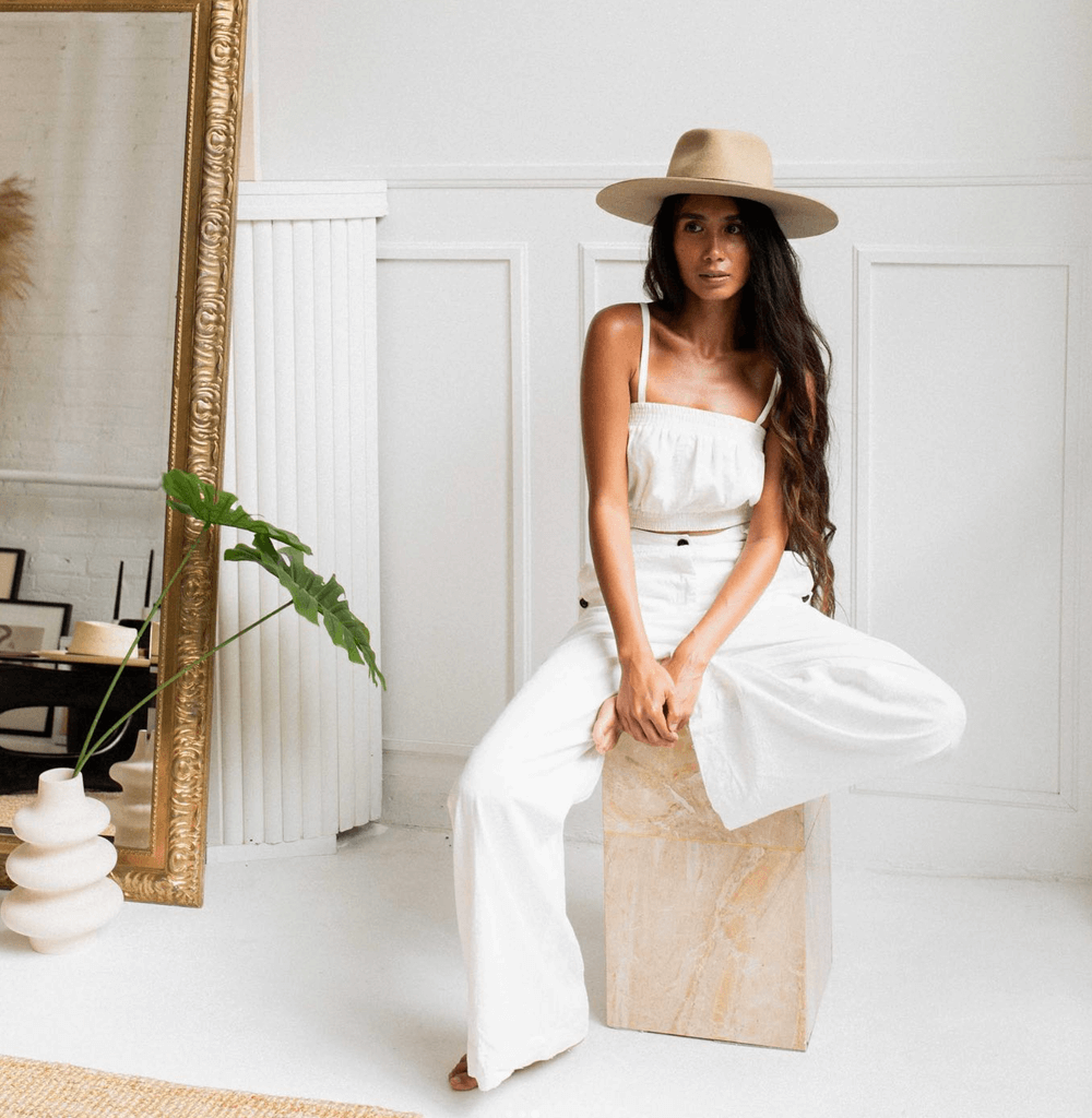 Verso Visual - A woman in white clothing and a large beige hat sitting on a wooden box in a minimalist room with white walls, a large mirror, and a potted plant.