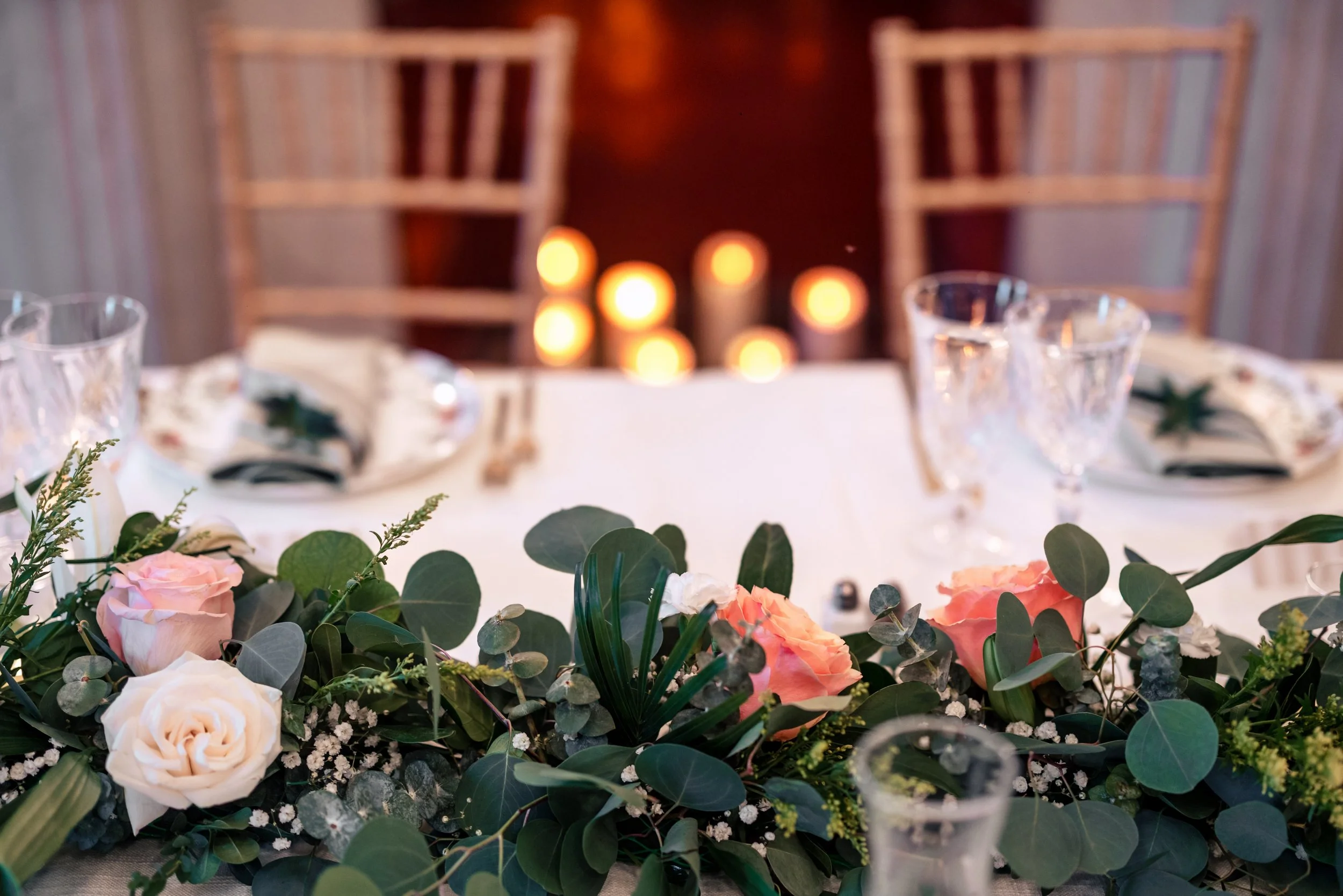 A beautifully decorated table with floral arrangements of pink and white roses, greenery, and baby's breath, set for a formal event, with candles in the background.
