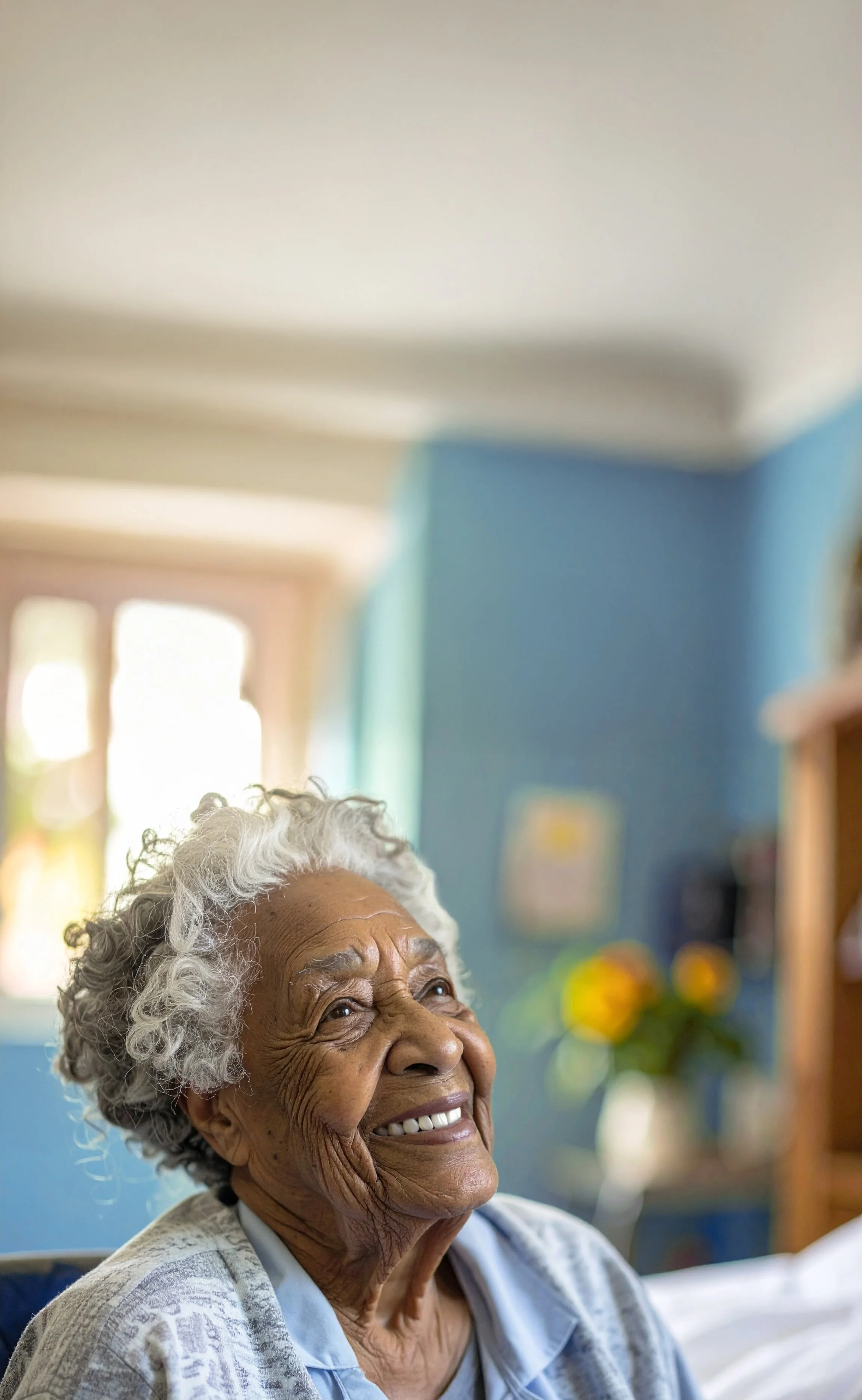 Smiling elderly woman with curly gray hair, sitting indoors in a sunlit room with blue walls, decorated with artwork and flowers in the background.