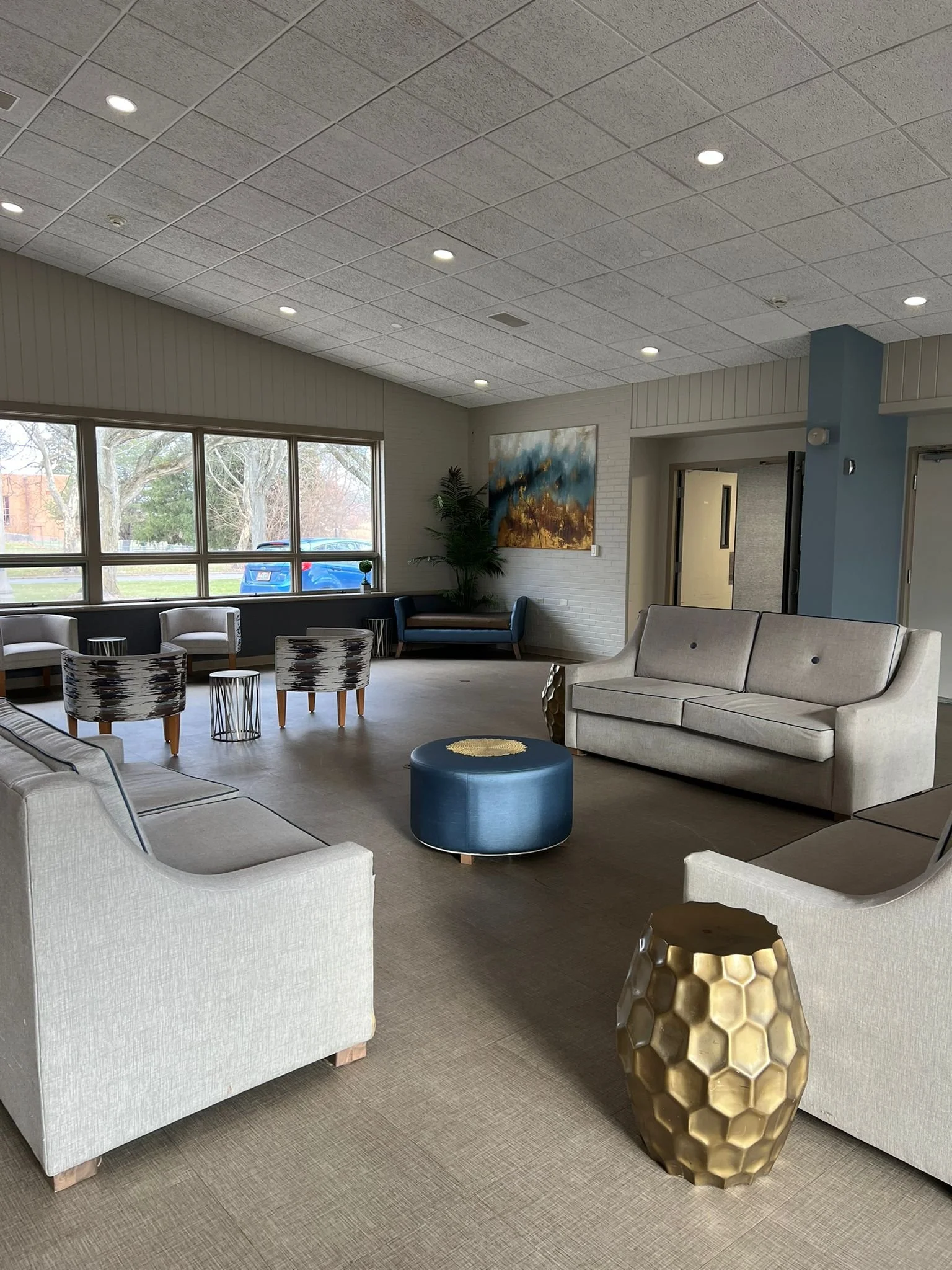A spacious modern lounge with beige sofas, patterned armchairs, a blue ottoman, and decorative gold vase. Large windows let in natural light, with art on the walls and a potted plant in the corner.