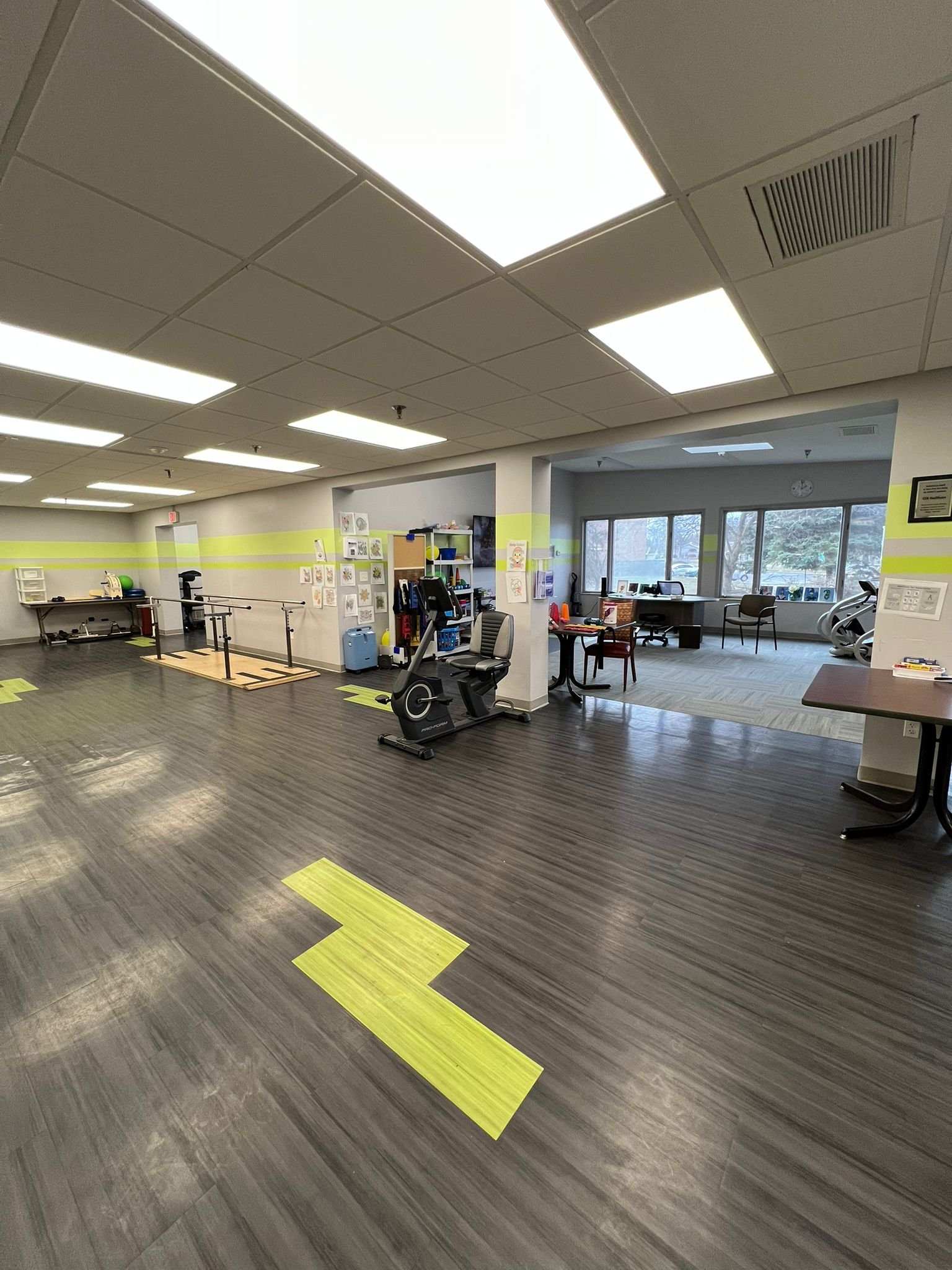 Empty physical therapy room with wooden flooring, exercise equipment, and large windows.
