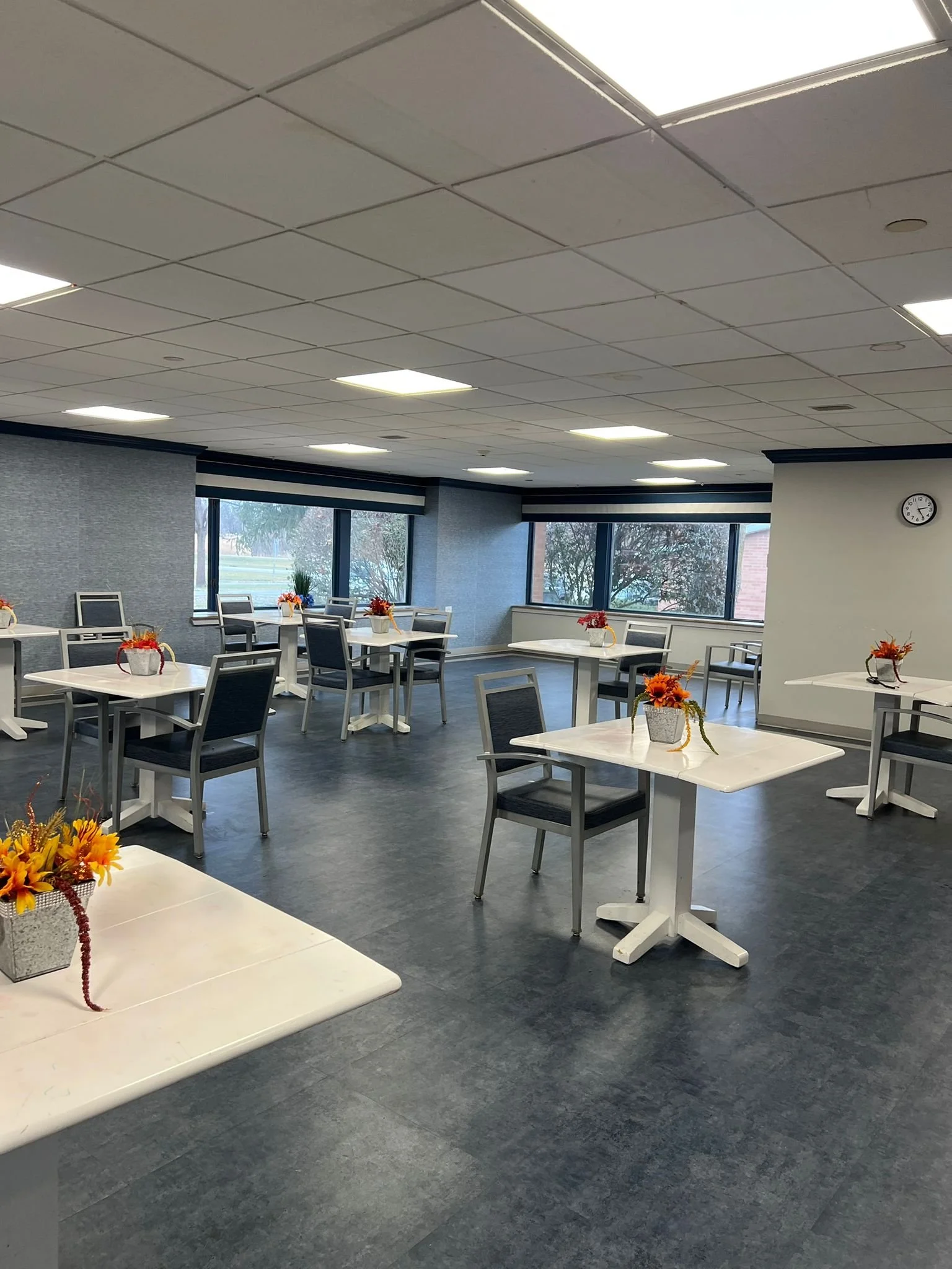 Empty dining area with white tables, black chairs, and fall-themed centerpieces, large windows, and a clock on the wall.