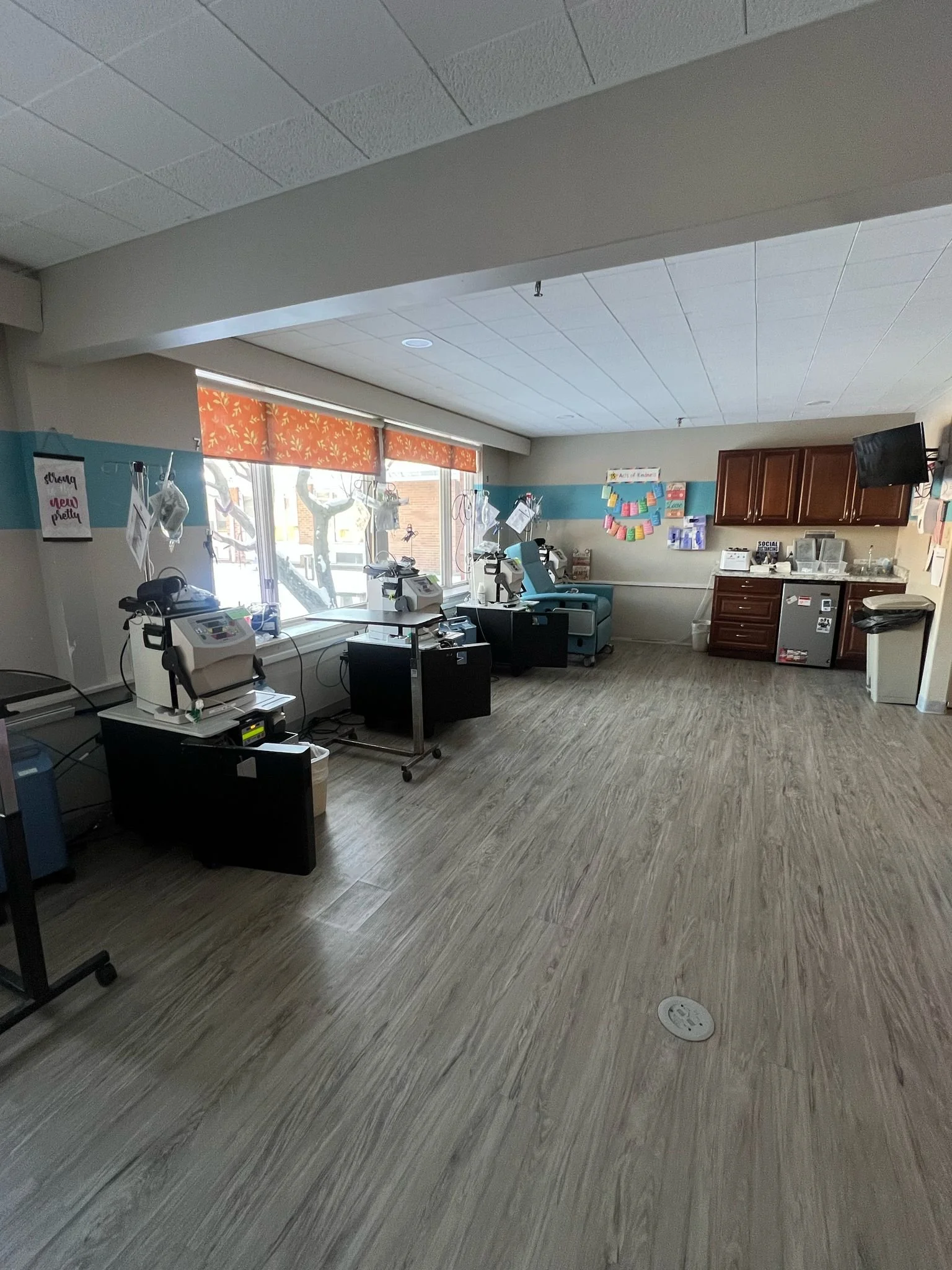 Empty hospital or medical treatment room with chairs, medical equipment, and medical supplies near windows, with a TV on the wall and colorful decorations.
