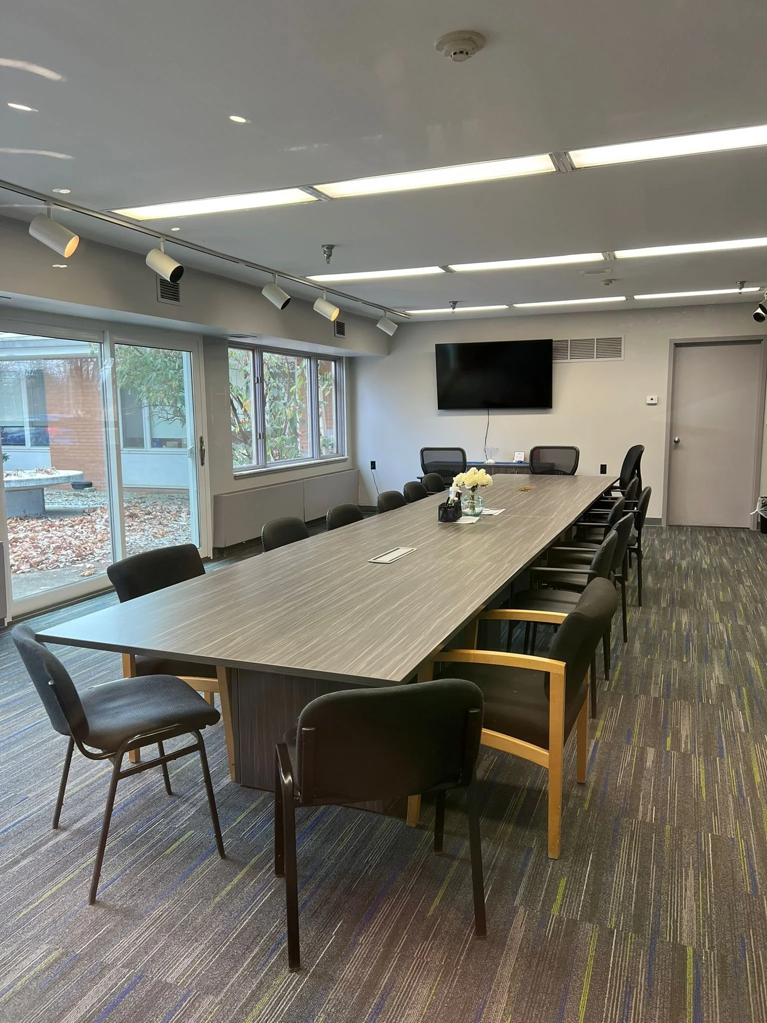 A modern conference room with a long rectangular table surrounded by black chairs, a large flat screen TV mounted on the wall, windows letting in natural light, and a door in the back corner.