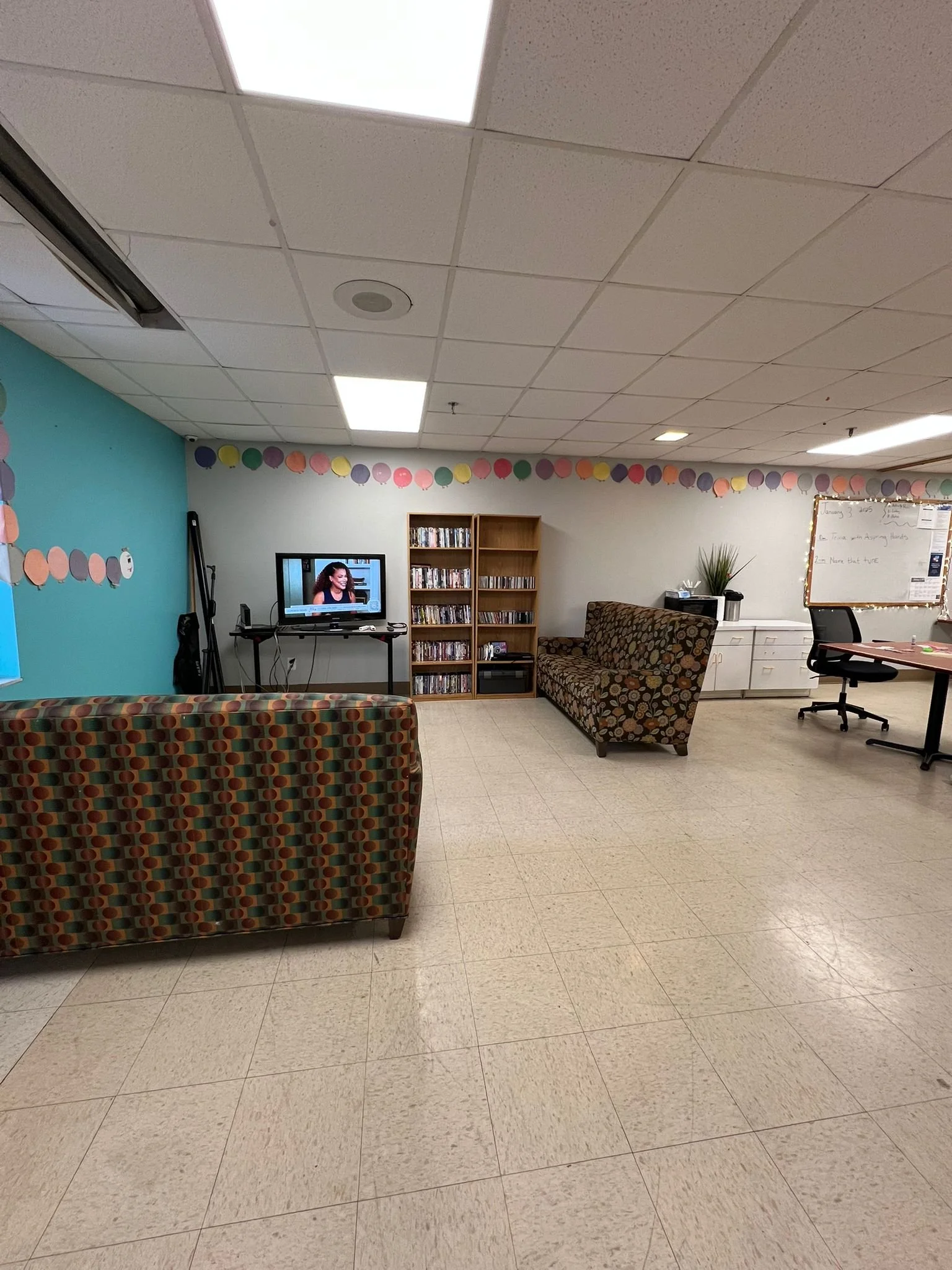 An empty room with a tiled floor and a white drop ceiling, decorated with multicolored paper circles along the upper walls. The room contains a patterned sofa facing a small television on a stand, a bookshelf nearby, and a desk with an office chair. A whiteboard is mounted on the wall.