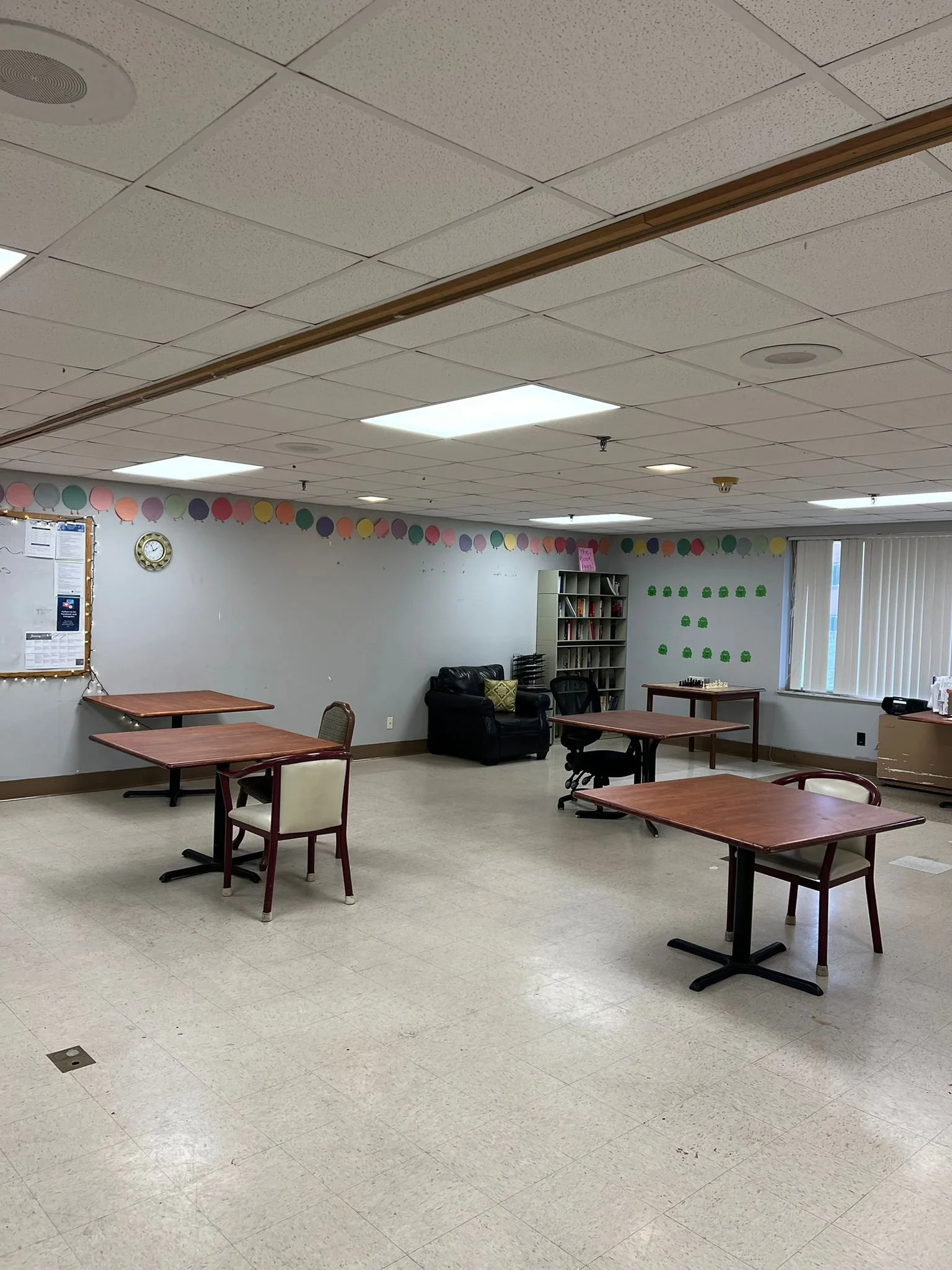 A classroom with a white ceiling, fluorescent lights, and tiled floor. There are three tables with chairs arranged around them, a black leather couch, a bookshelf filled with books, a clock on the wall, colorful balloon garlands hung along the wall, and a window with vertical blinds.