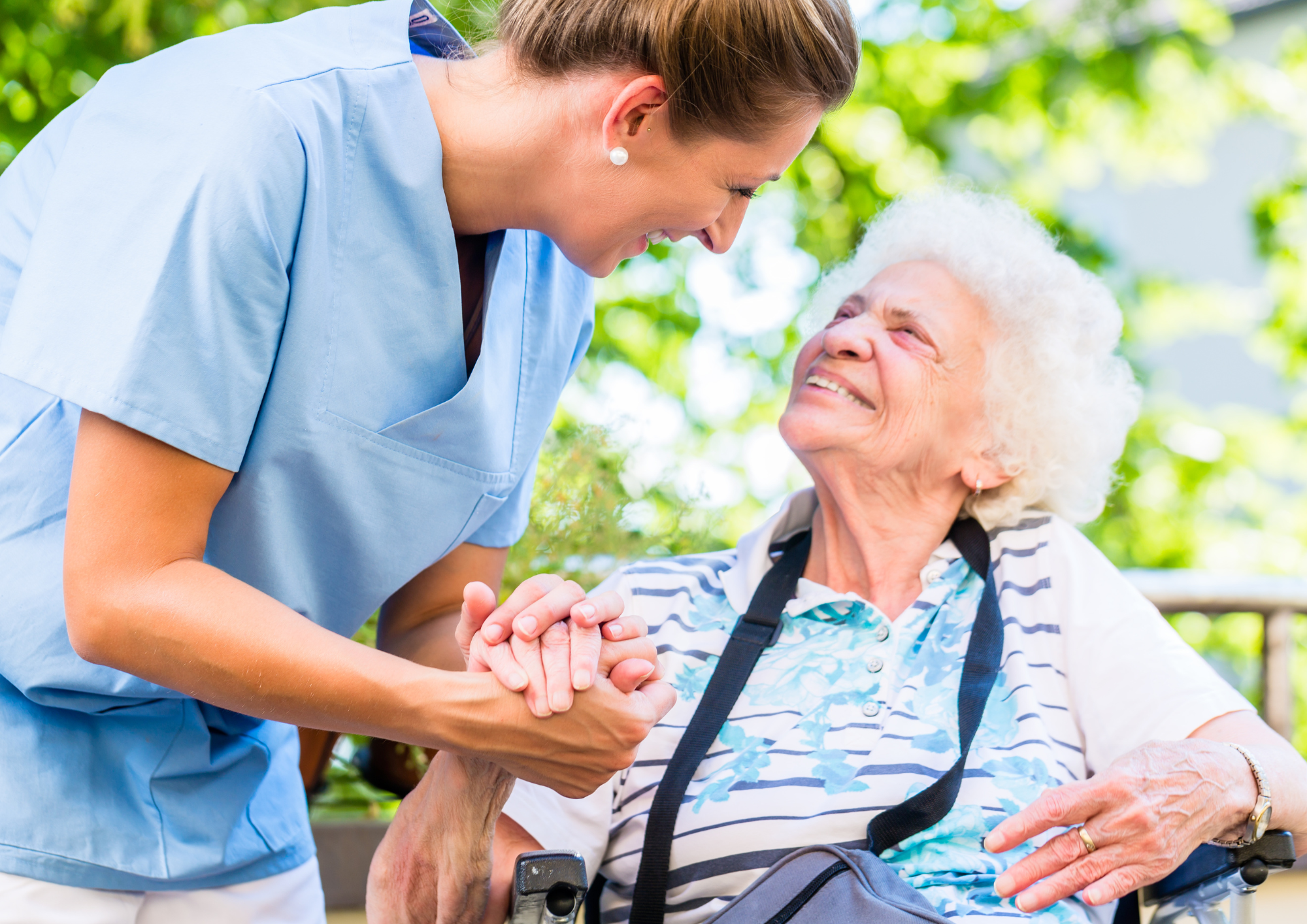 A young nurse or caregiver smiling at an elderly woman in a wheelchair outdoors, holding her hands and sharing a joyful moment.