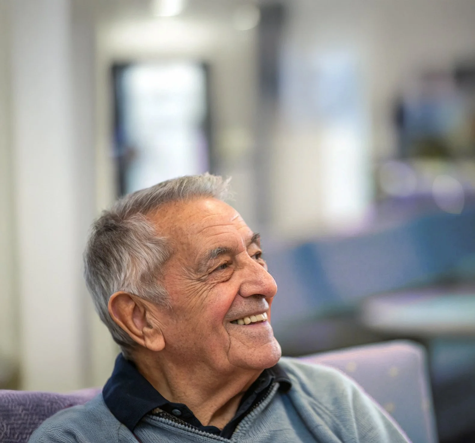 An elderly man with gray hair and a mustache, smiling and looking to his left, sitting in a chair in an indoor setting.
