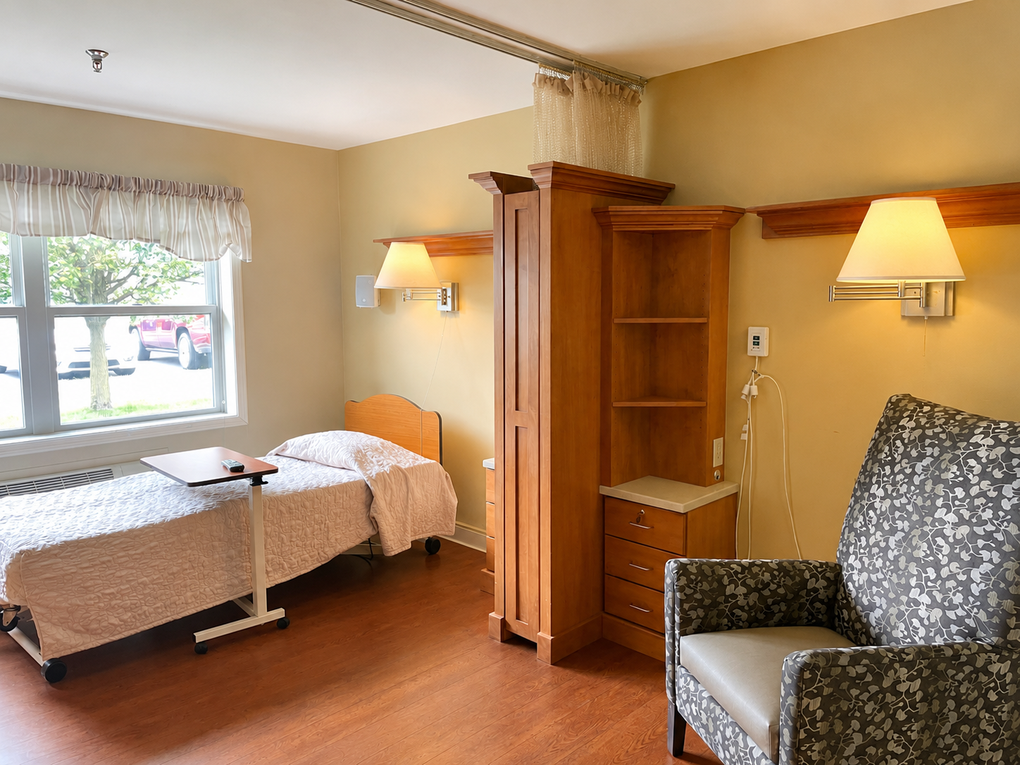 Hospital room with a bed, window, wooden furniture, armchair, and wall lamps.