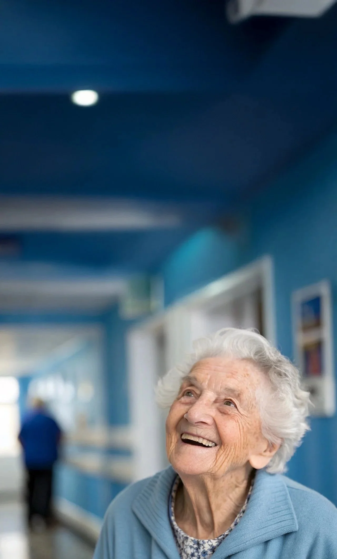 An elderly woman with white hair smiling and looking up, wearing a blue sweater and patterned shirt, in a brightly lit room with blue walls and framed pictures.