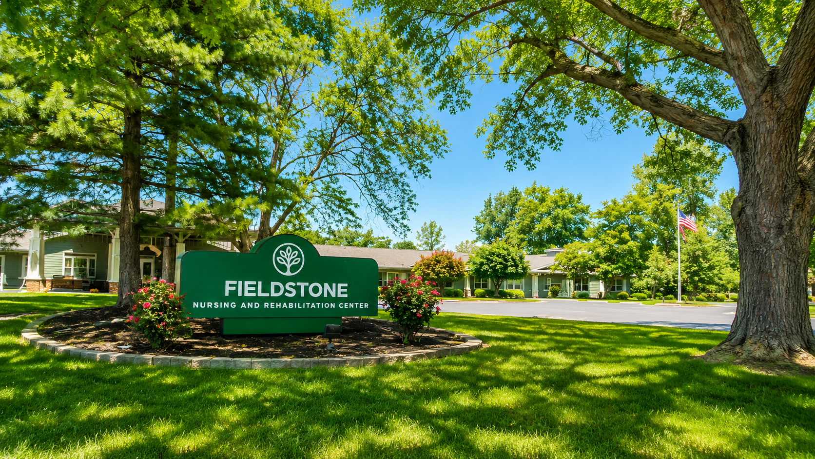 Exterior view of the Fieldstone Nursing and Rehabilitation Center with a large green sign in front, surrounded by flowers and greenery, under a bright blue sky with trees and a flagpole with the American flag.