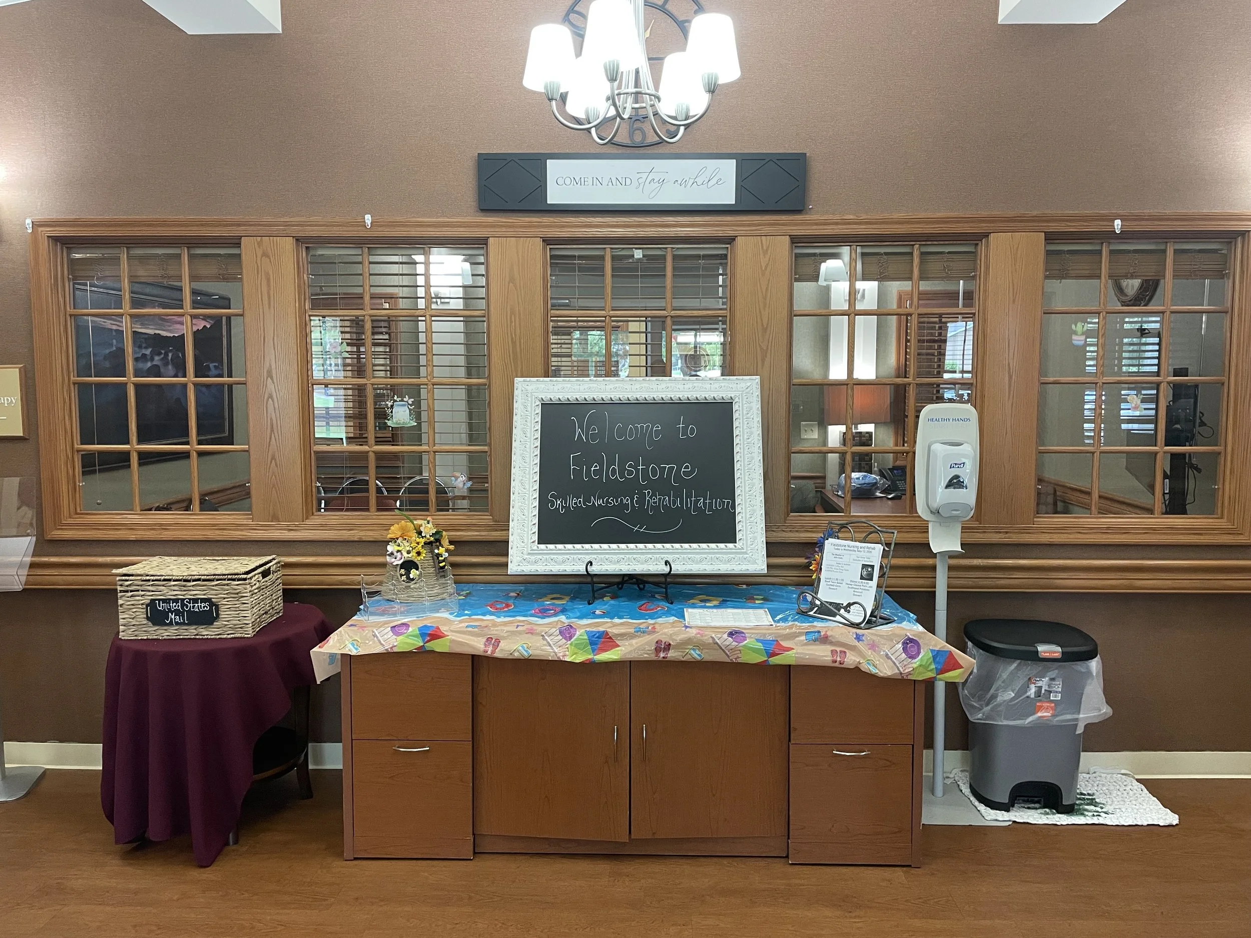 Reception desk with a chalkboard sign welcoming visitors to Fieldstone Skilled Nursing and Rehabilitation, decorated with a bouquet of yellow flowers, and a table with a colorful umbrella-themed tablecloth in front of a wooden partition with glass windows. A chandelier hangs above, and a sign reads, 'Come in and stay awhile.' Hand sanitizer and mail basket are also visible.