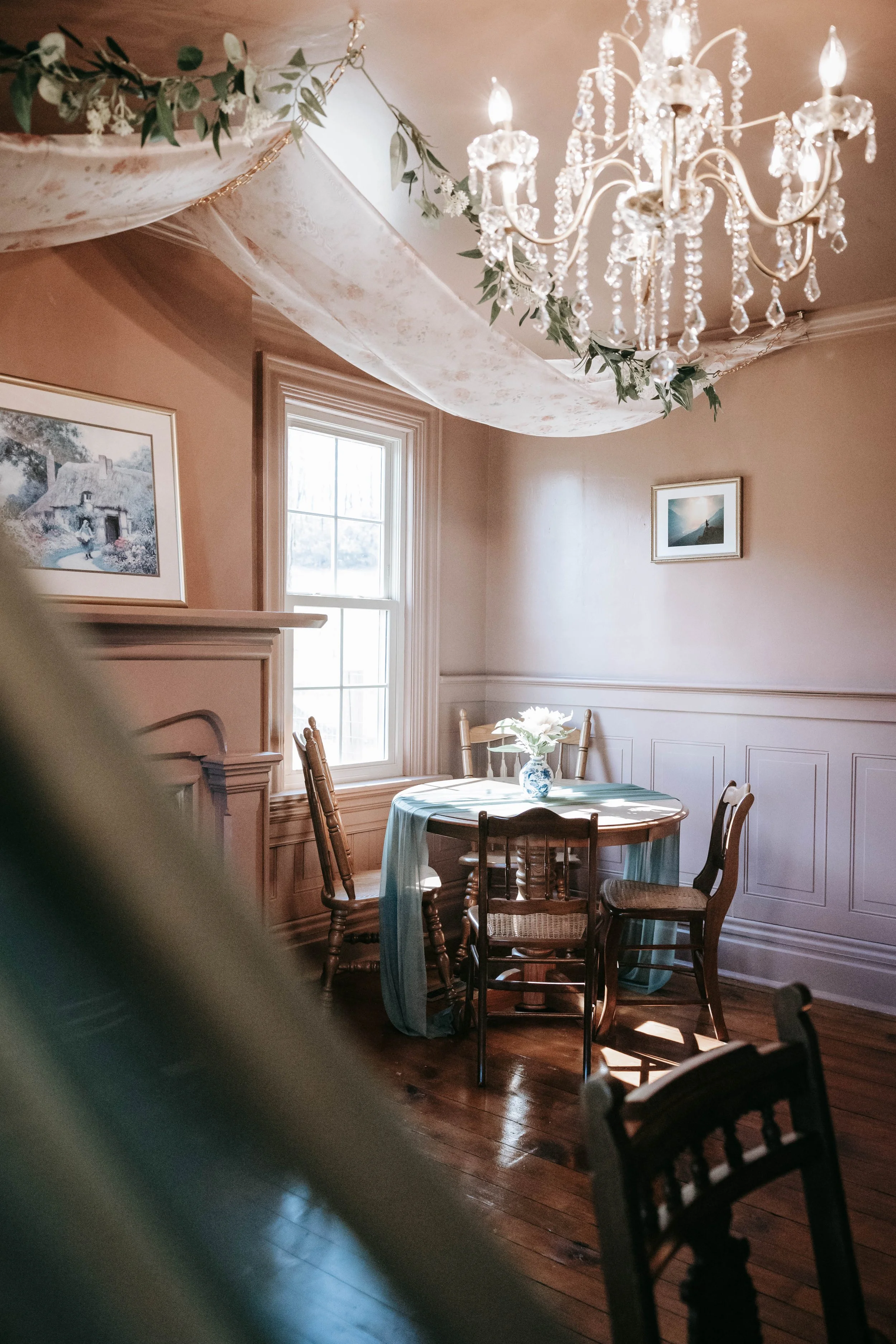 A cozy dining room with a wooden round table, six wooden chairs, a vase with white flowers, a window letting in natural light, a chandelier hanging from the ceiling, and framed pictures on the walls.
