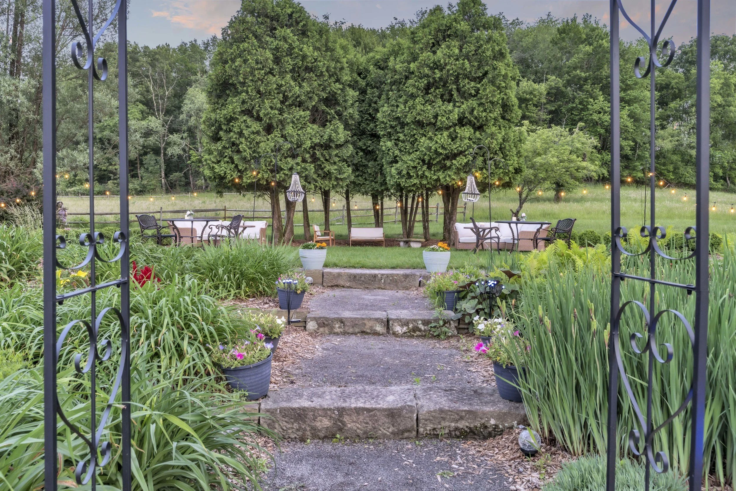 View through an open wrought iron gate into a garden setting with a stone pathway, potted plants, lush green foliage, and outdoor seating area with tables and chairs, surrounded by string lights and tall trees in the background.