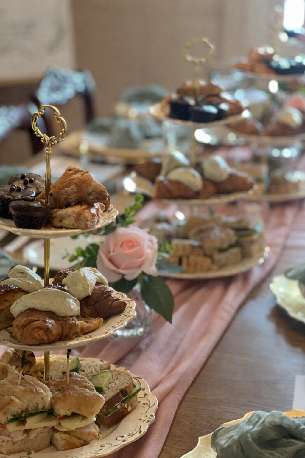 A tiered serving tray with assorted baked goods such as croissants, scones, cookies, and finger sandwiches, decorated with a pink rose and greenery, set on a table with a pink tablecloth.
