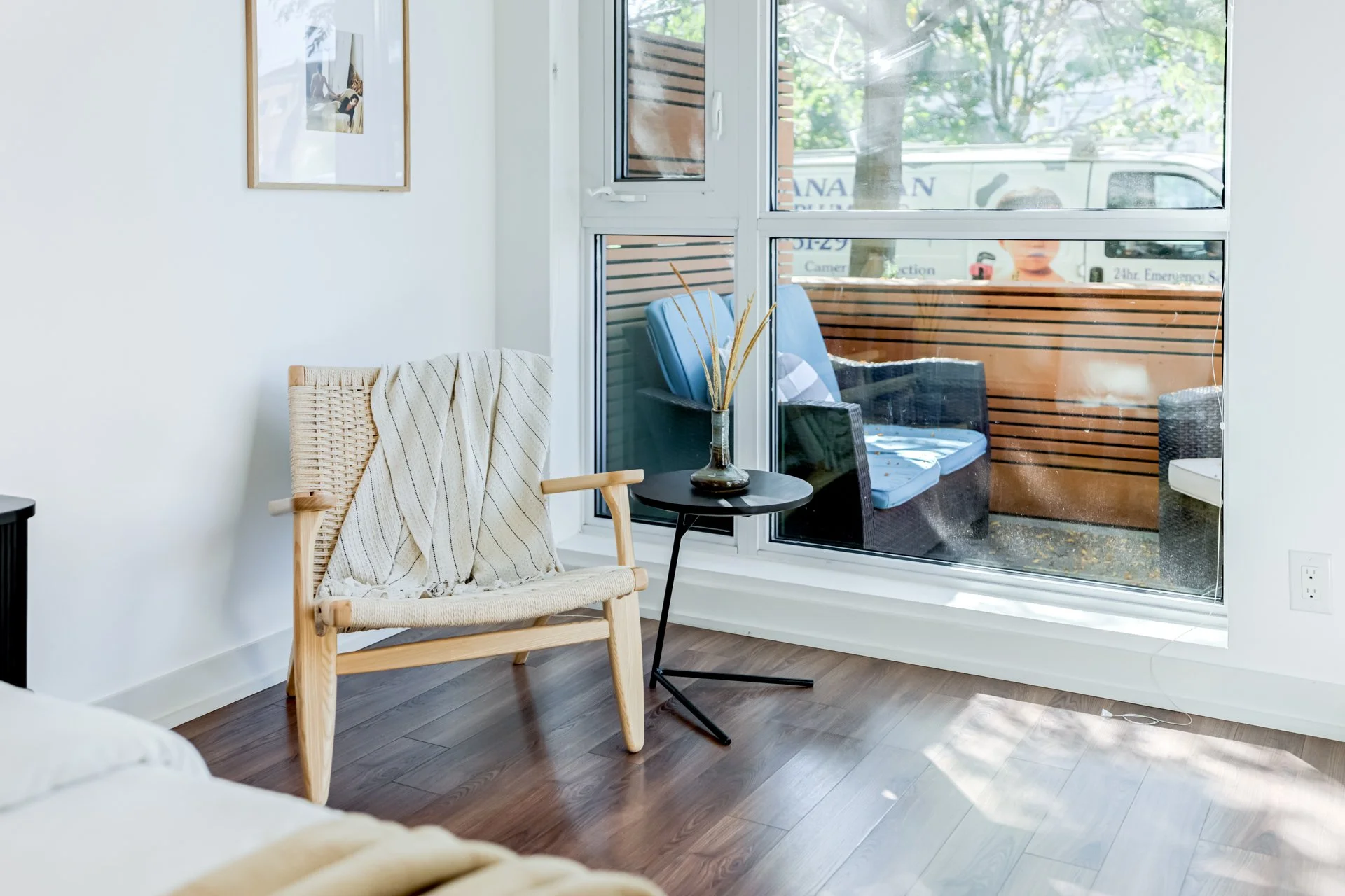 A cozy interior with a woven chair, a small black side table with a glass vase and dried decorative grasses, and a large window showing a balcony with outdoor chairs and a view of trees outside.
