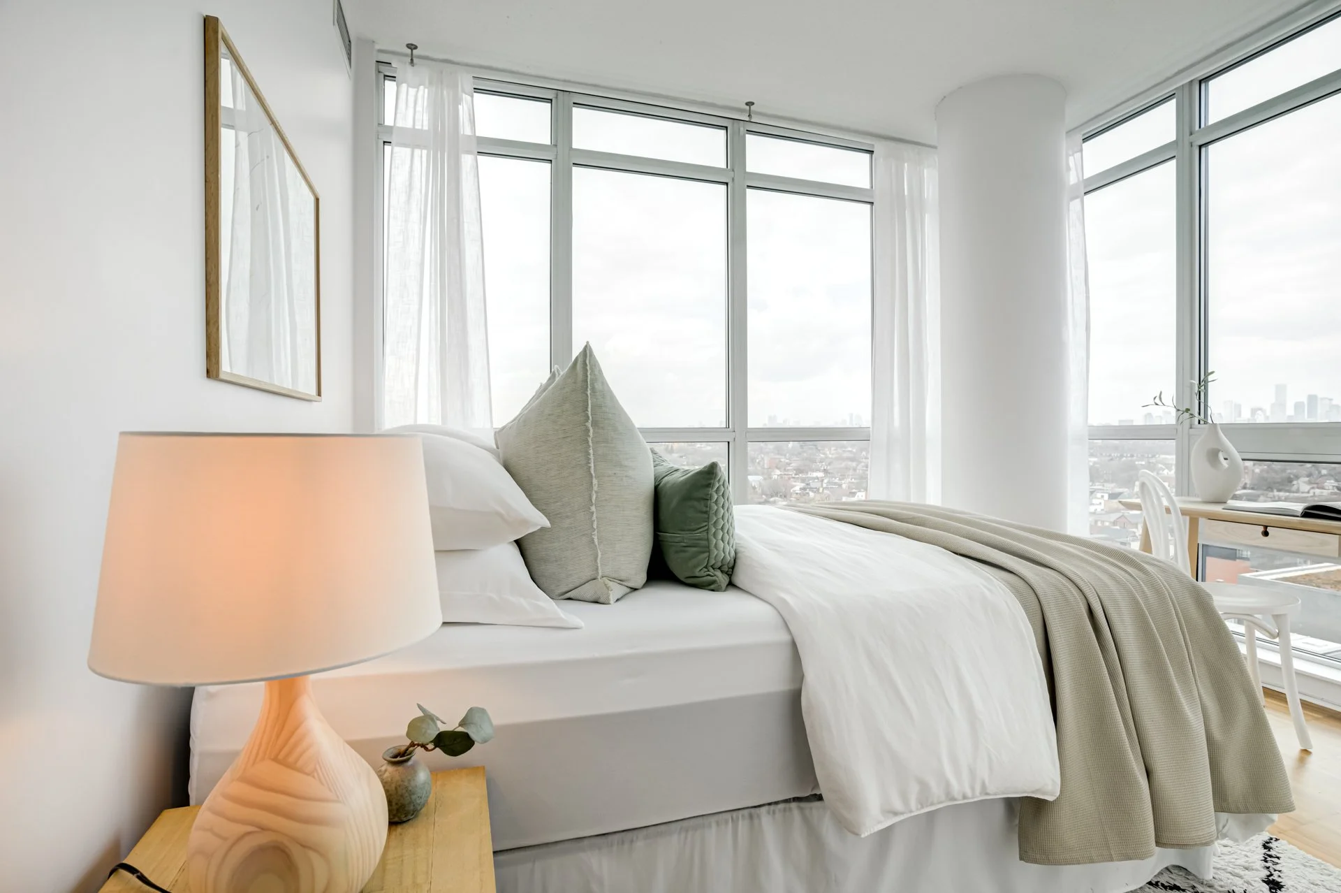Bright modern bedroom with white bedding, decorative pillows, and large windows with sheer curtains showing cityscape view.