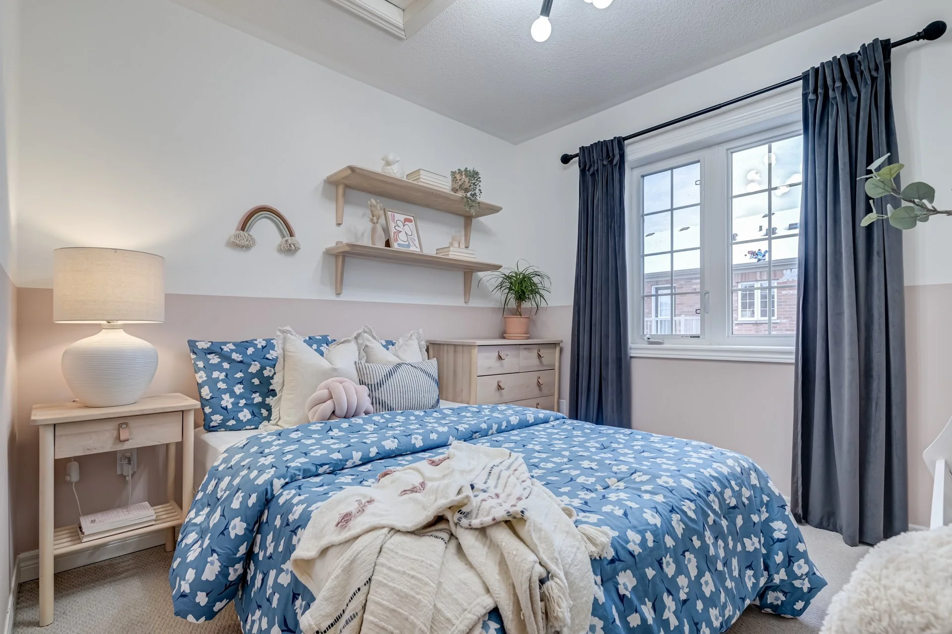 Cozy bedroom with blue and white floral bedding, pillows, a nightstand with lamp, wooden dresser, floating shelves with decor, a window with dark blue curtains, and potted plants.