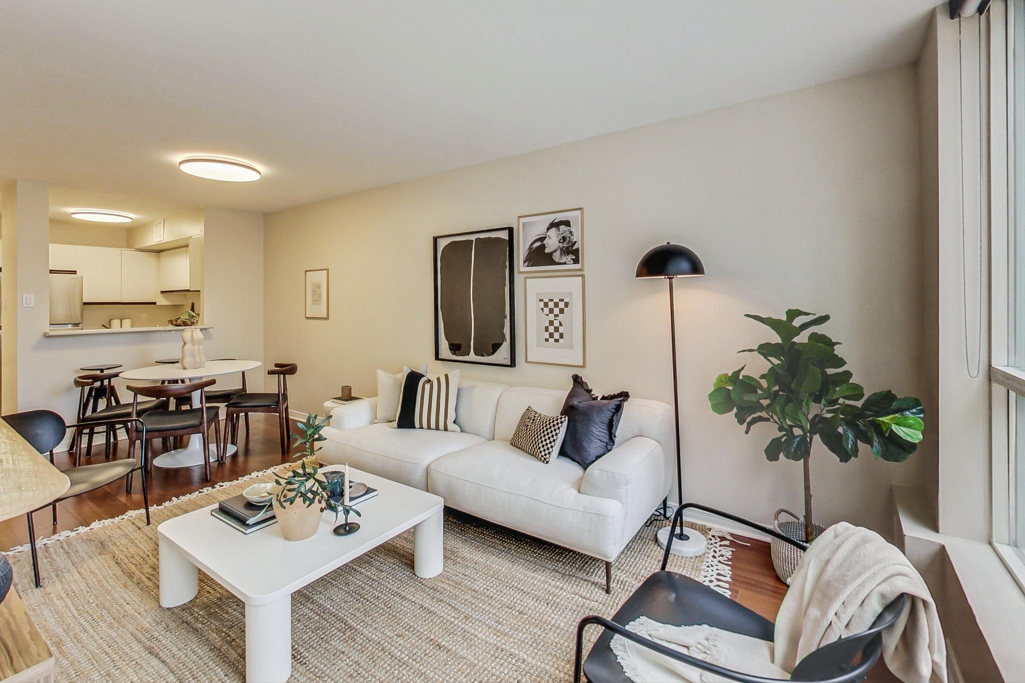Modern living room with white sofa, black and patterned throw pillows, black floor lamp, large green plant, coffee table with decorative items, beige area rug, and wall art, opening to kitchen area.