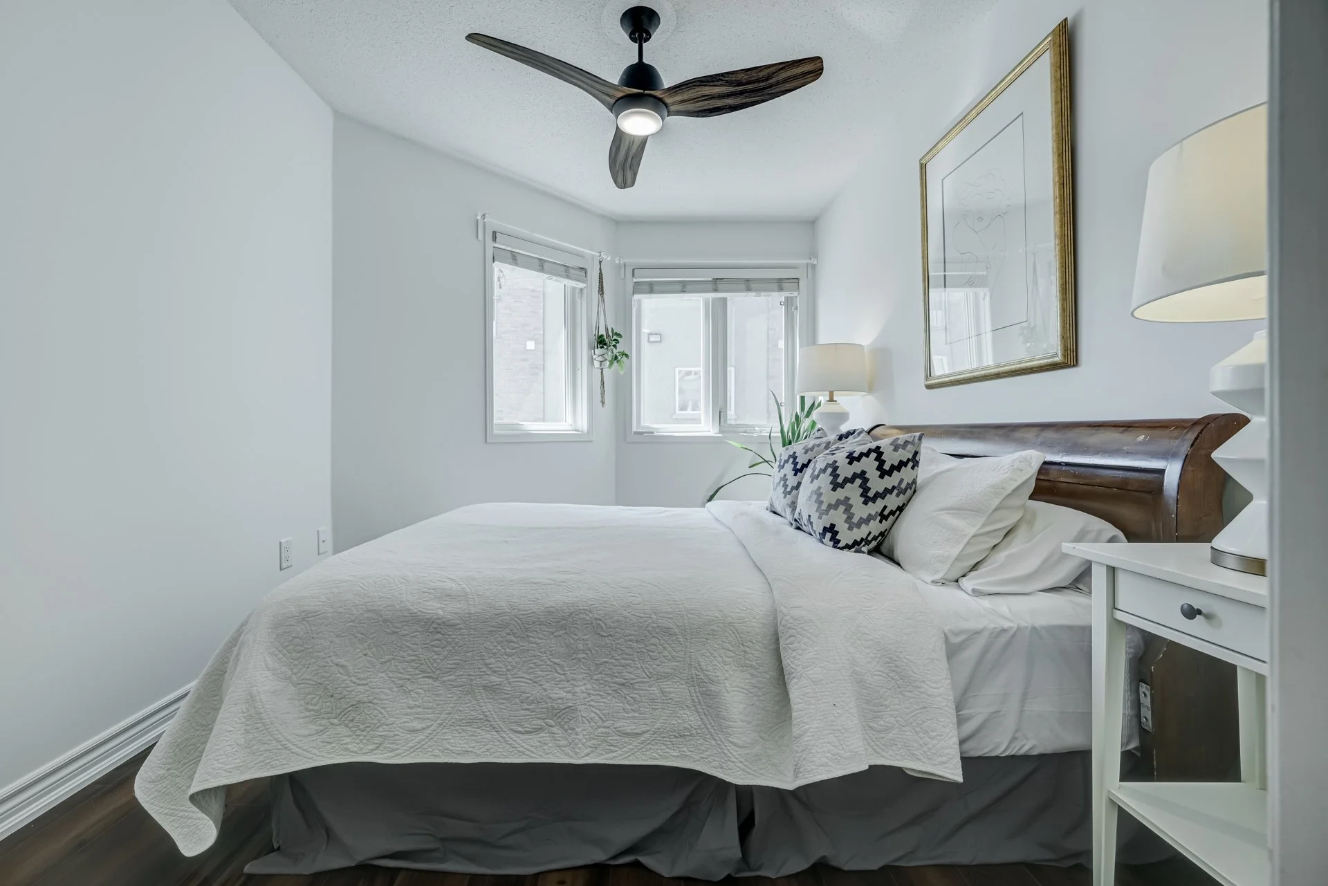 A bedroom with a white bedspread, pillows, nightstand, lamps, a ceiling fan, potted plant, framed art, and three windows with blinds, in a bright, modern style.