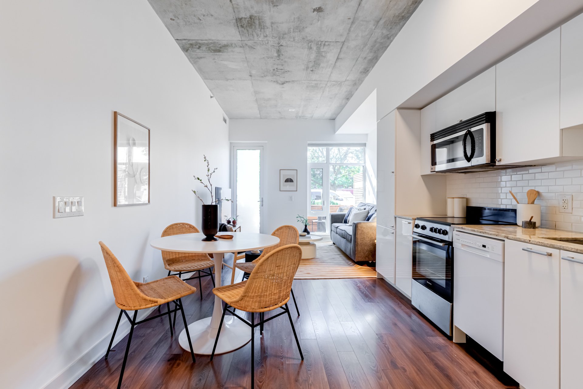Open-concept kitchen and dining area with white cabinets, black appliances, a gray couch at the far end, and large windows letting in natural light. There is a white dining table with four wicker chairs, and hardwood floors.