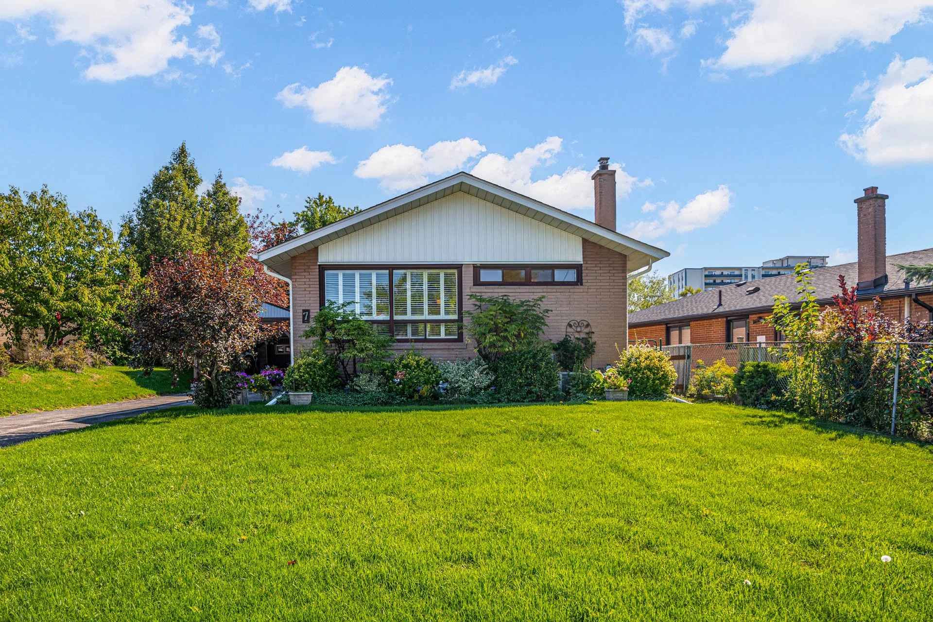 A single-story house with a white gable roof, brick walls on the front, and a large front window with blinds. The house is surrounded by a well-maintained green lawn, various bushes, and trees, with blossoming flowers in pots. The background shows a blue sky with scattered white clouds.
