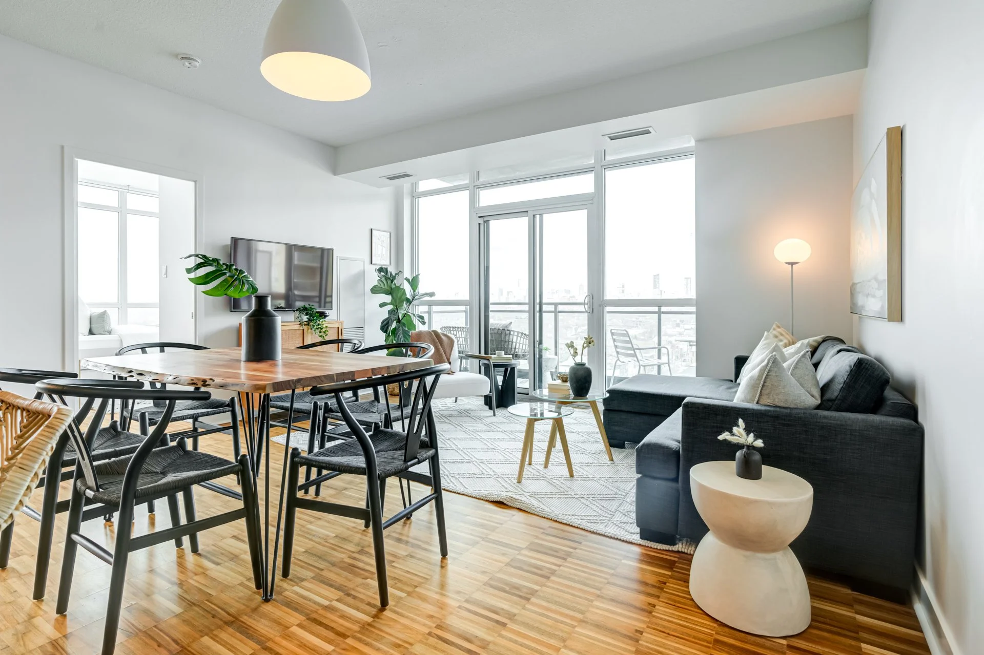 Modern living room with large floor-to-ceiling windows, black sofa, coffee table, flat-screen TV, and dining area with a wooden table and black chairs.