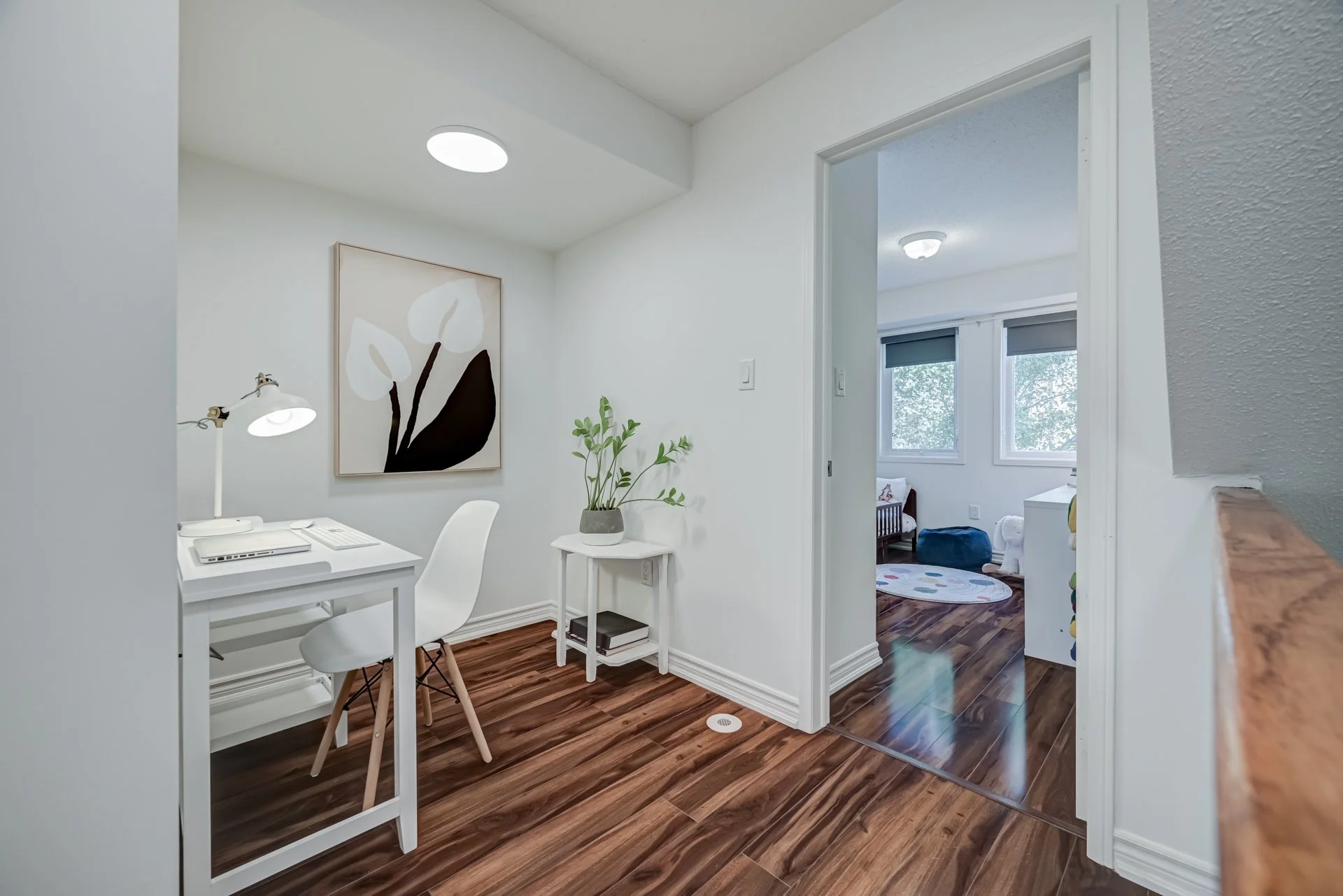 A bright, white home interior with a small workspace that has a white desk, a white chair, a desk lamp, and a plant on a side table. There is a large framed abstract artwork on the wall behind the desk. The room has brown wood flooring and is adjacen