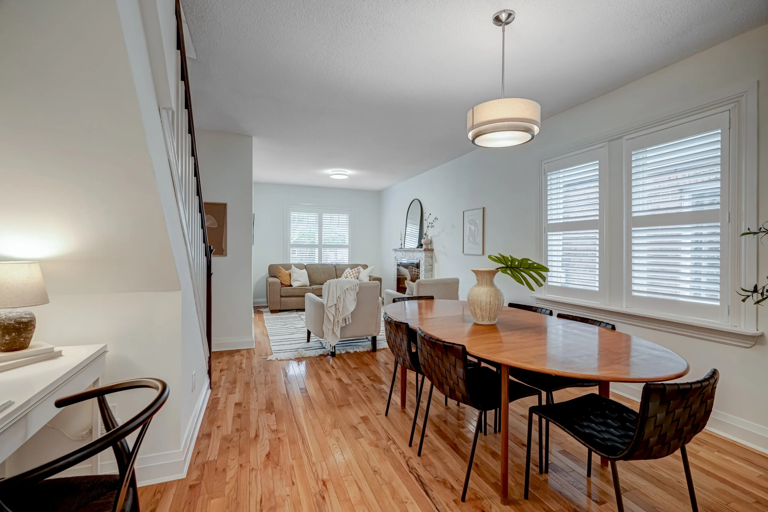 Living and dining room with hardwood floors, white walls, large windows with white blinds, a beige sofa, white armchairs, a fireplace with a mirror above it, a round wooden dining table with black chairs, a large vase with a green leaf as decor, and 