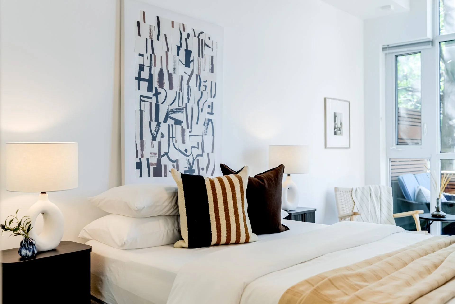 A cozy bedroom with a large bed, white bedding, and decorative pillows, flanked by two nightstands with white lamps, and a sitting area with chairs and a window view outside.