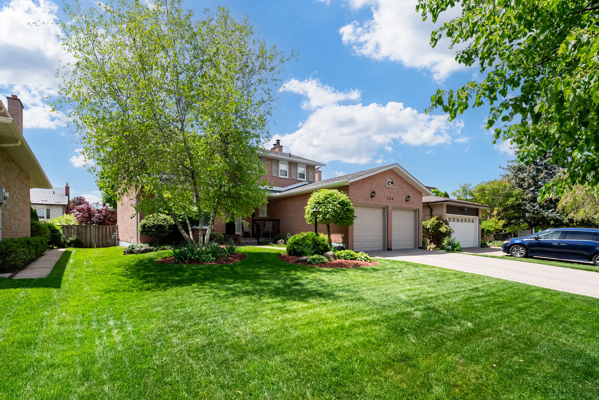 A suburban house with a well-kept lawn, trees, and a driveway with parked car under a partly cloudy sky.
