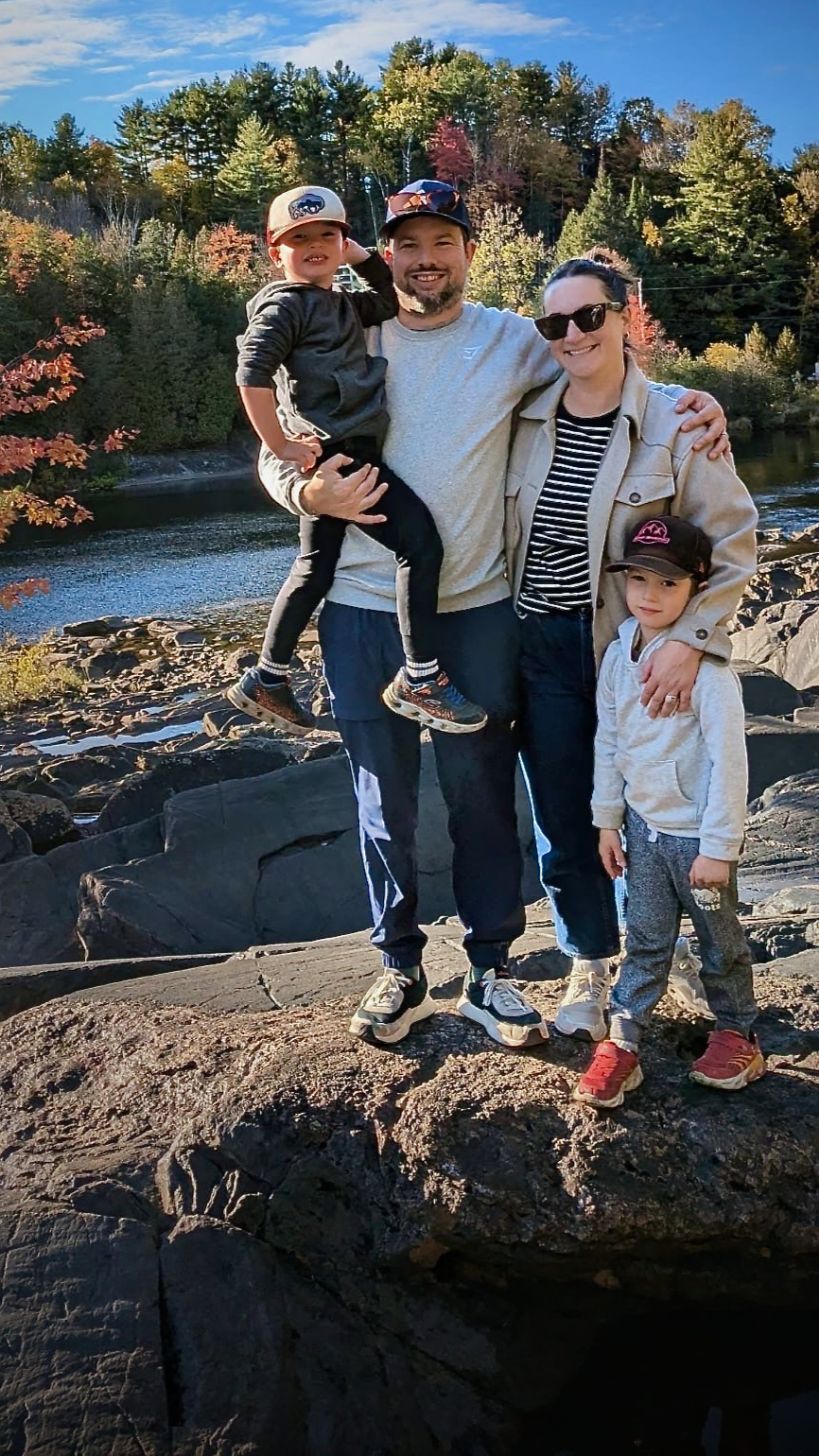 A family of four stands on rocks near a body of water with a wooded area and a blue sky in the background. The father holds a young boy, and a woman and a girl stand beside him, all smiling.