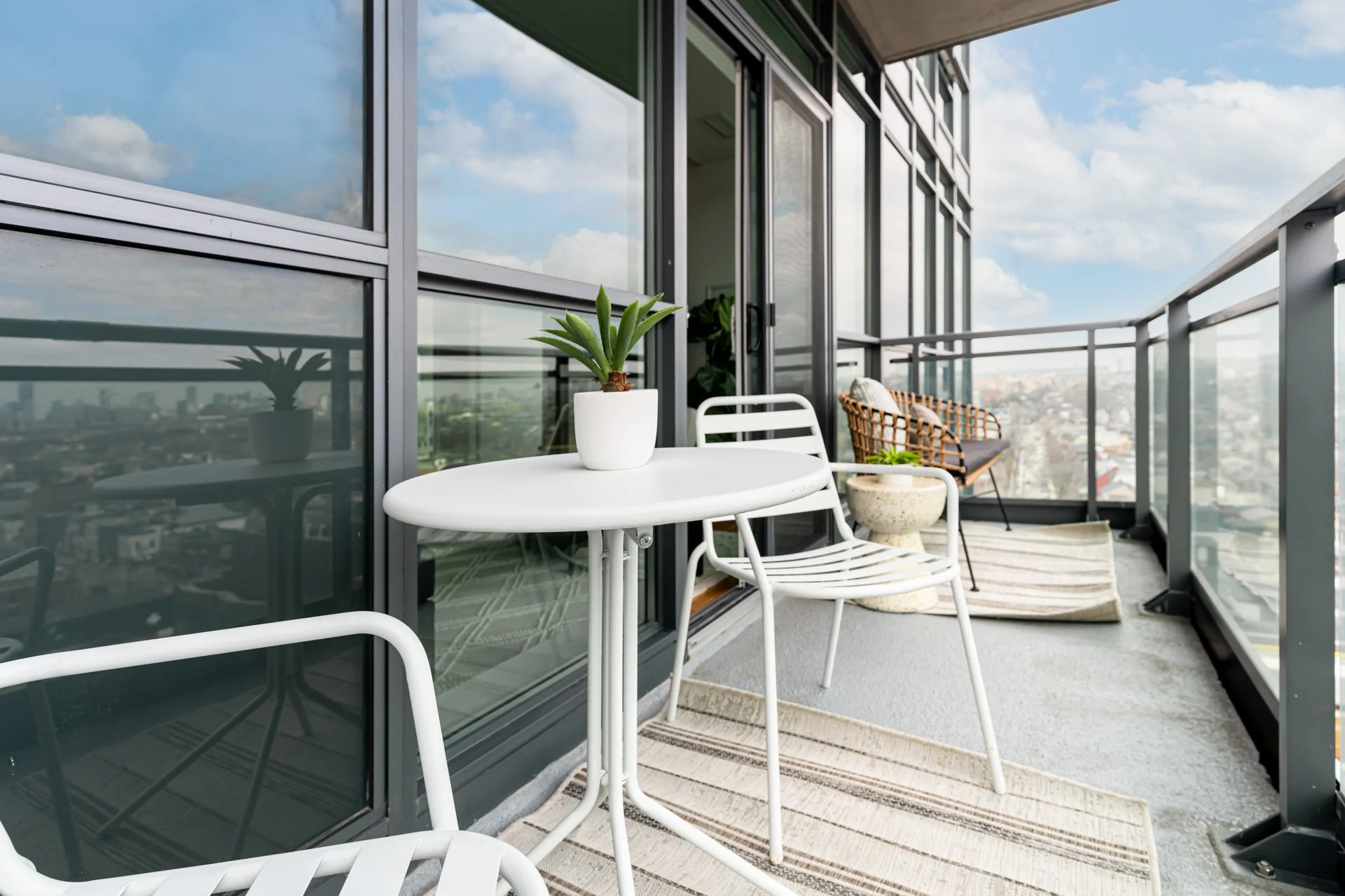 Balcony with white table, chairs, potted plant, rug, railing, city view, blue sky, clouds