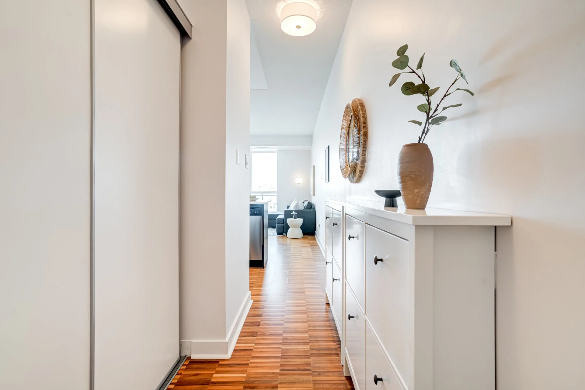 Interior of an apartment hallway with wooden flooring, a white console table decorated with a large clay vase with greenery, a black bowl, and a woven wall mirror. In the background, a living room with a gray sectional sofa and a window letting in na