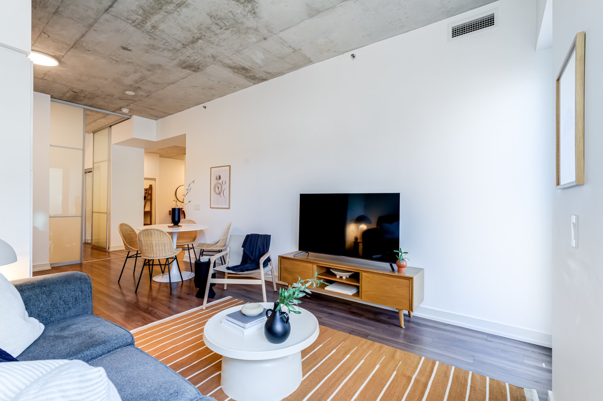 Modern living room with a gray sofa, wooden coffee table, flat-screen TV on a wooden TV stand, and a dining area with a round table and four chairs.