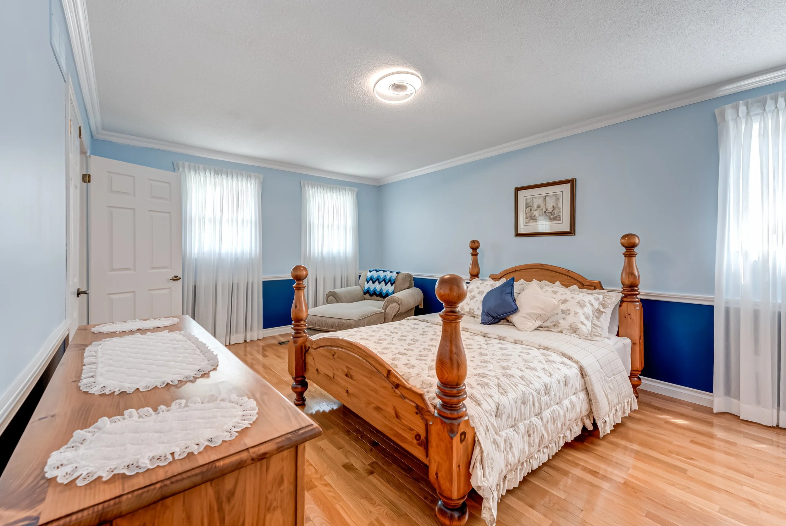 A cozy bedroom with light blue walls, a wooden bed frame with bedding and pillows, white curtains on windows, and a comfortable chair with a matching ottoman near the wall.